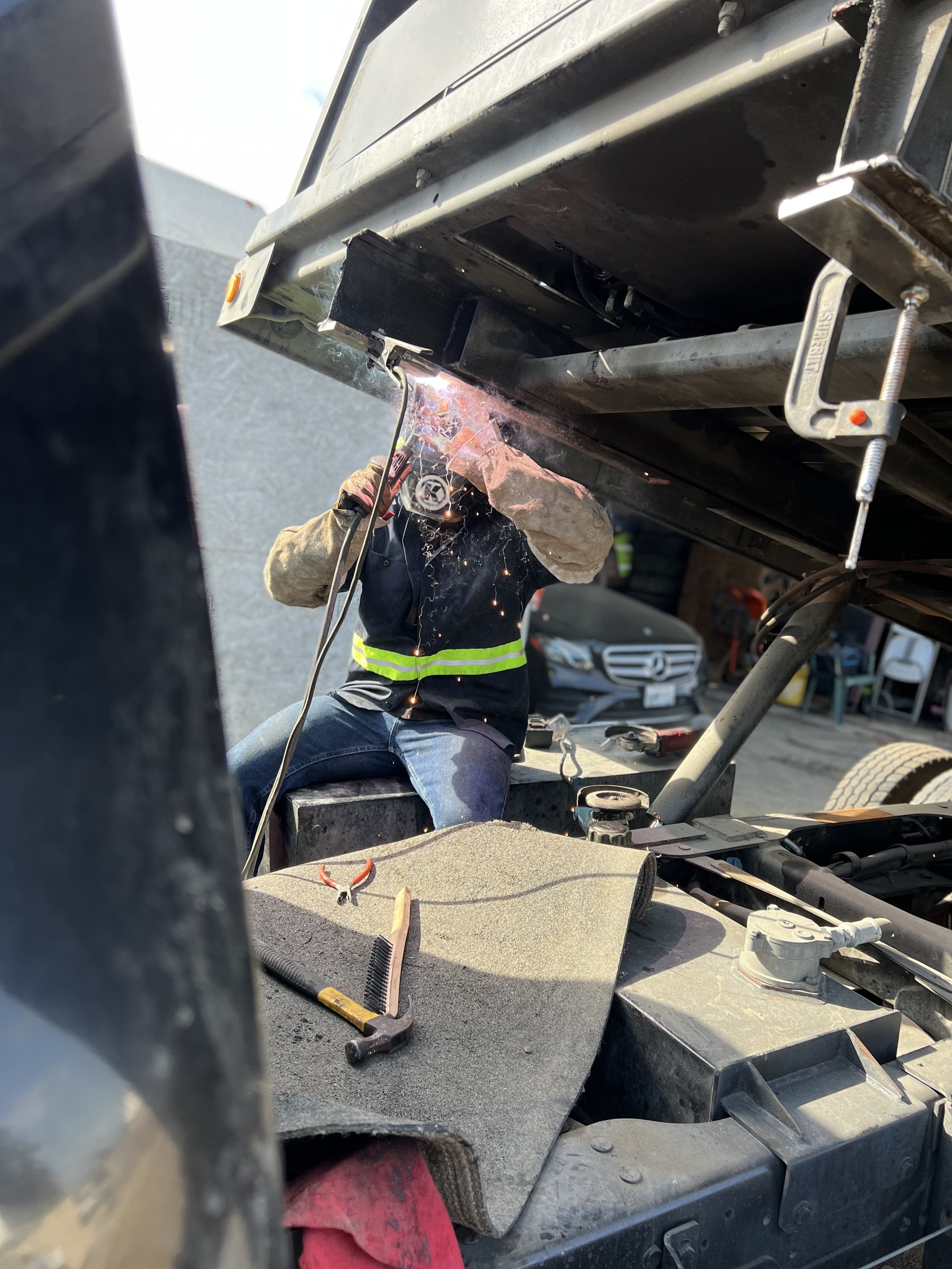 A person welding underneath a large vehicle or trailer, with sparks flying. Various tools and equipment are visible around.