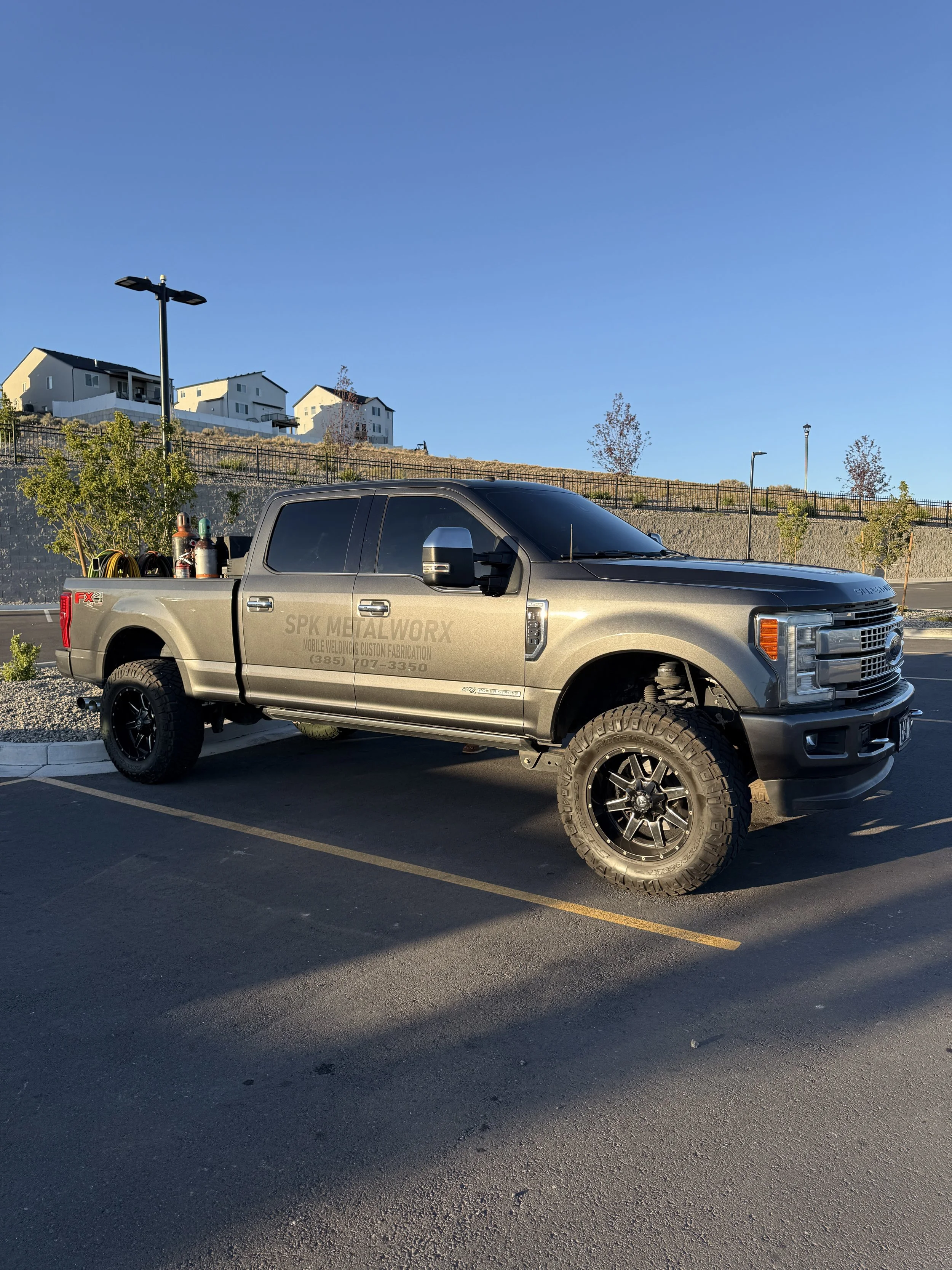 A silver pickup truck parked in a parking lot with several tools and equipment in the truck bed, under a clear blue sky with buildings on a hillside in the background.