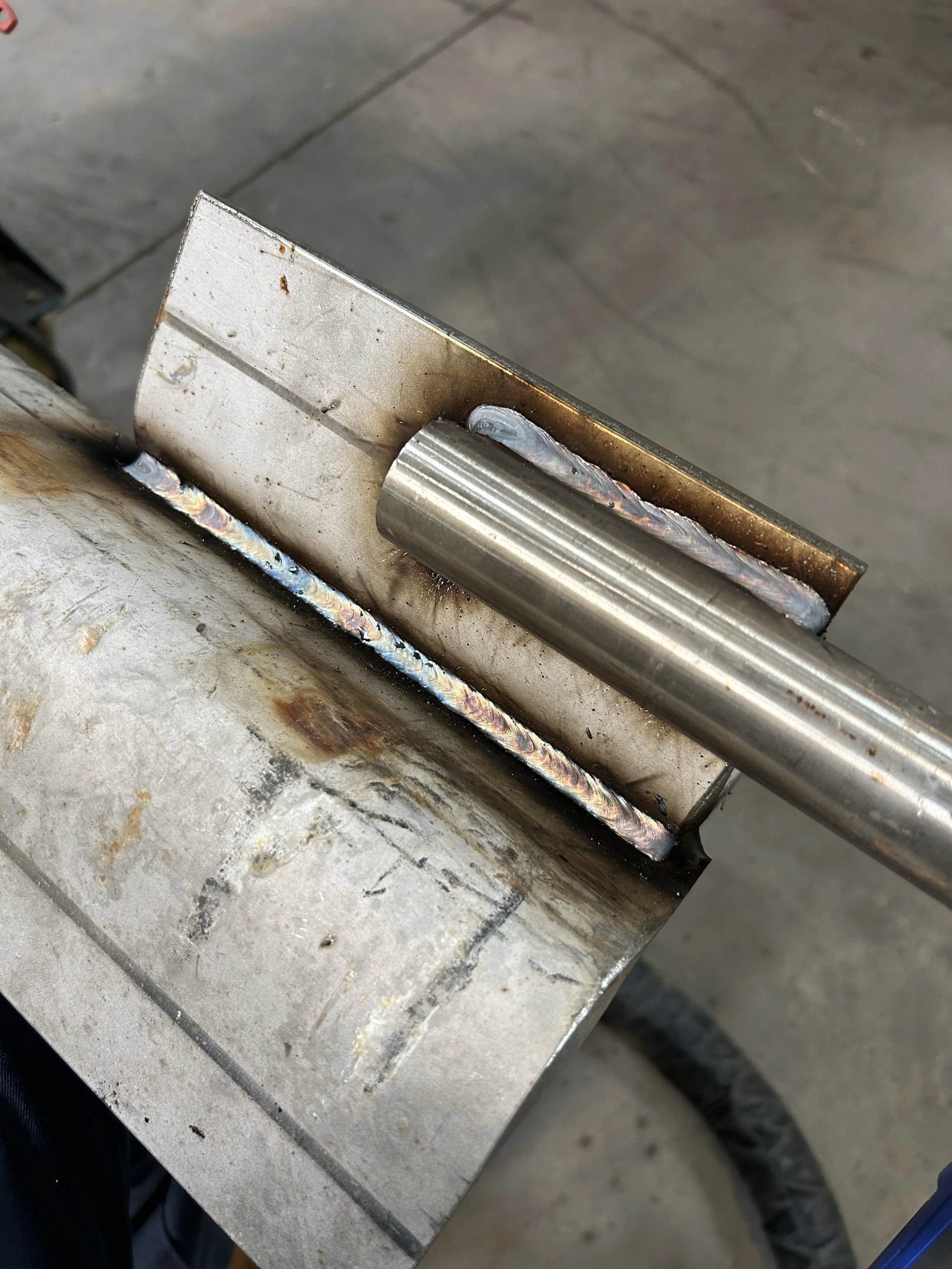 Close-up view of a metal welding joint on a metal piece, showing welding beads and some discoloration, on a flat metal surface in an industrial setting.