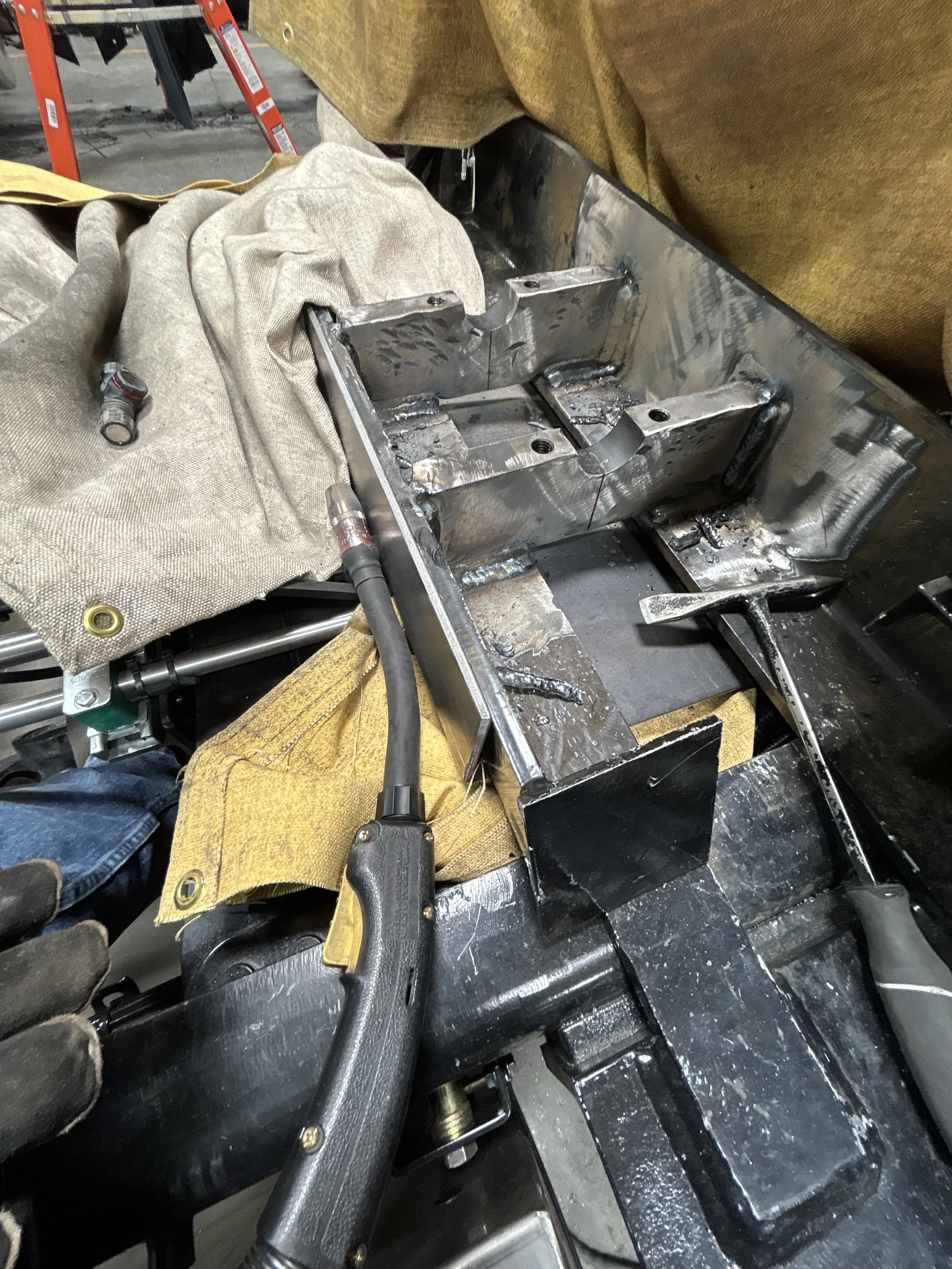 Close-up of a metalwork project with a person welding or grinding, surrounded by welding tools, gloves, and protective towels, with a ladder and workshop background.