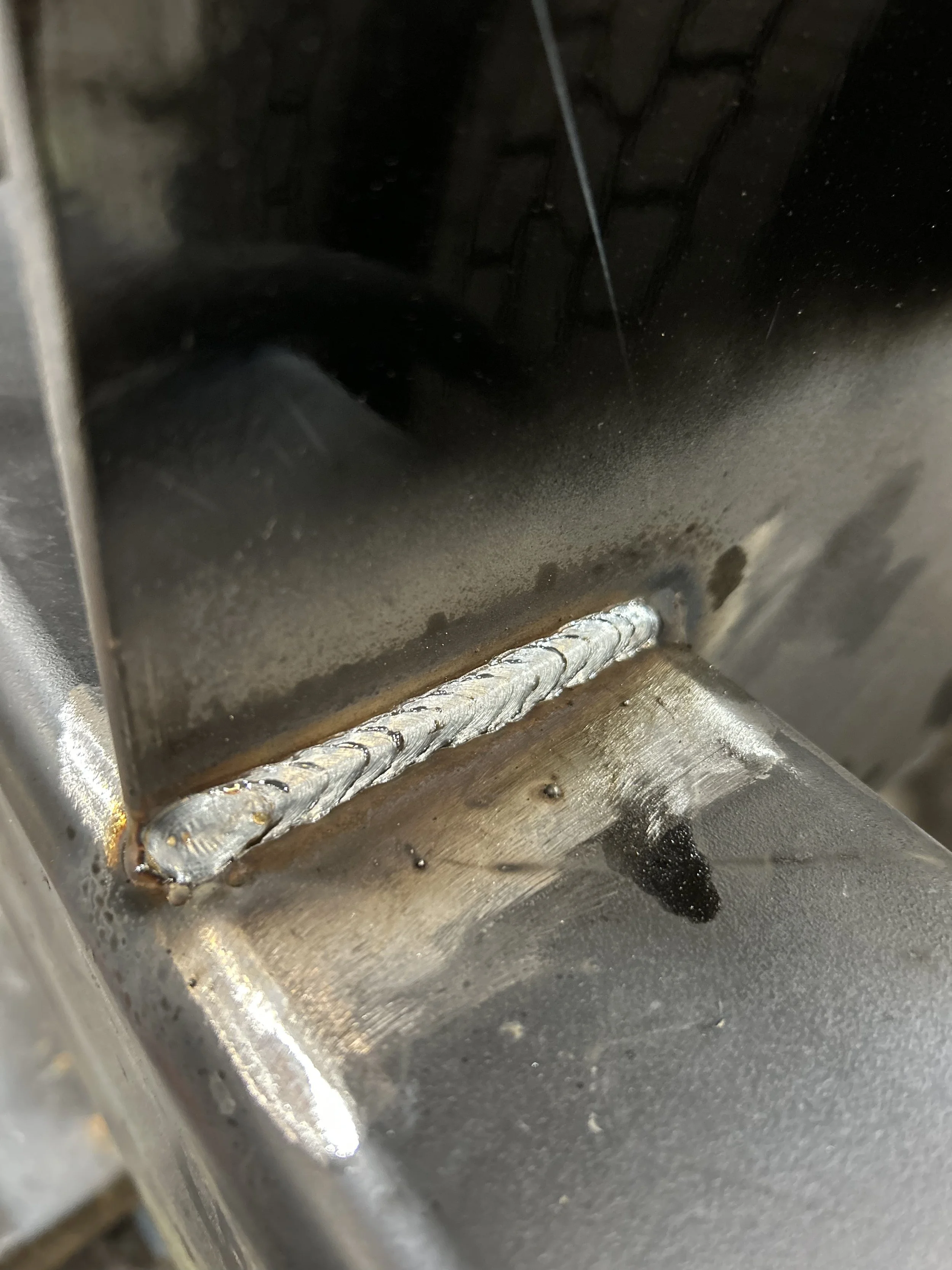 Close-up of a metal surface with a welded joint, showing shiny weld beads and some discoloration and residue.