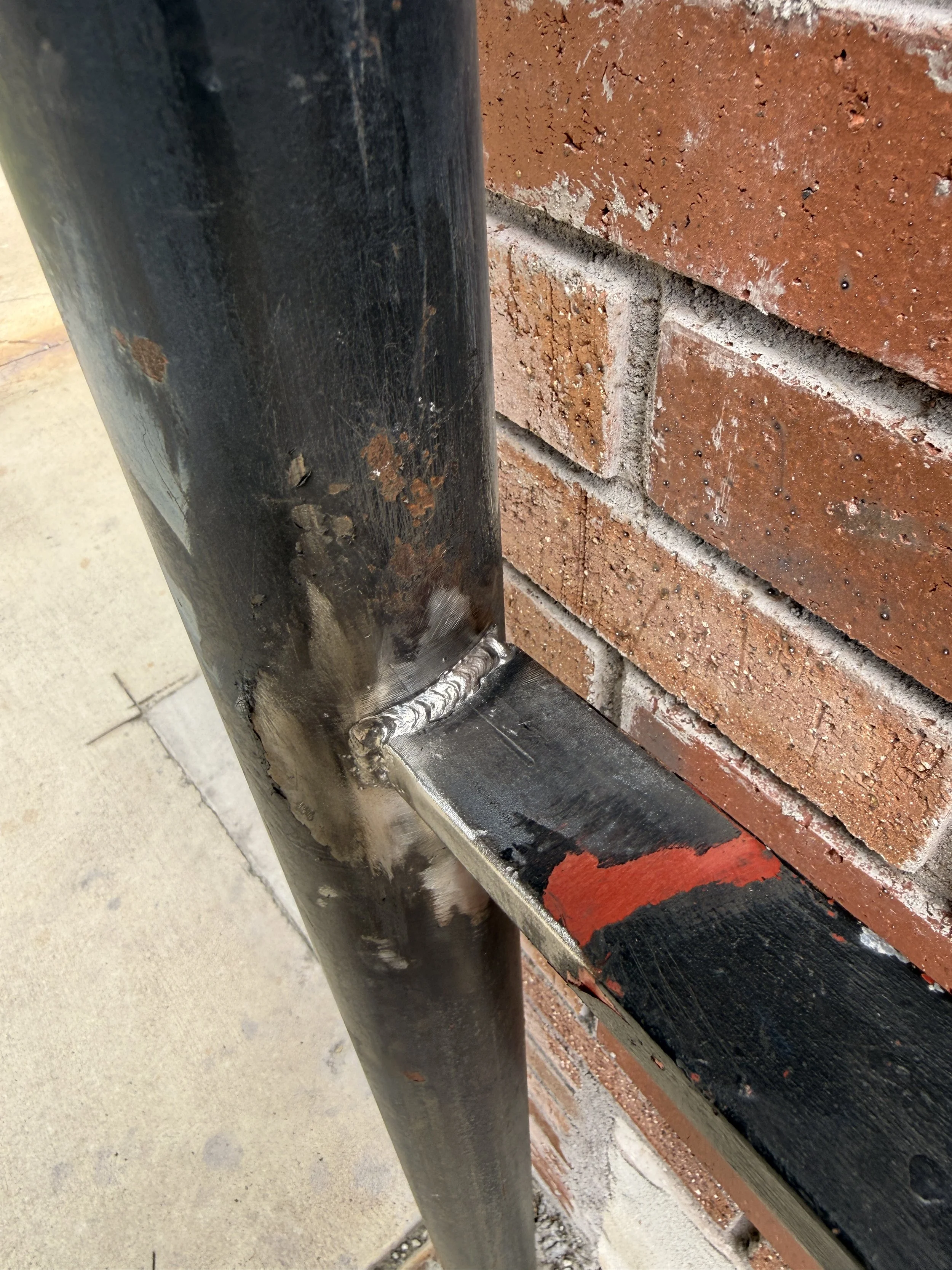 Close-up of a weathered black handrail attached to a red brick wall, with visible welds at the connection point.