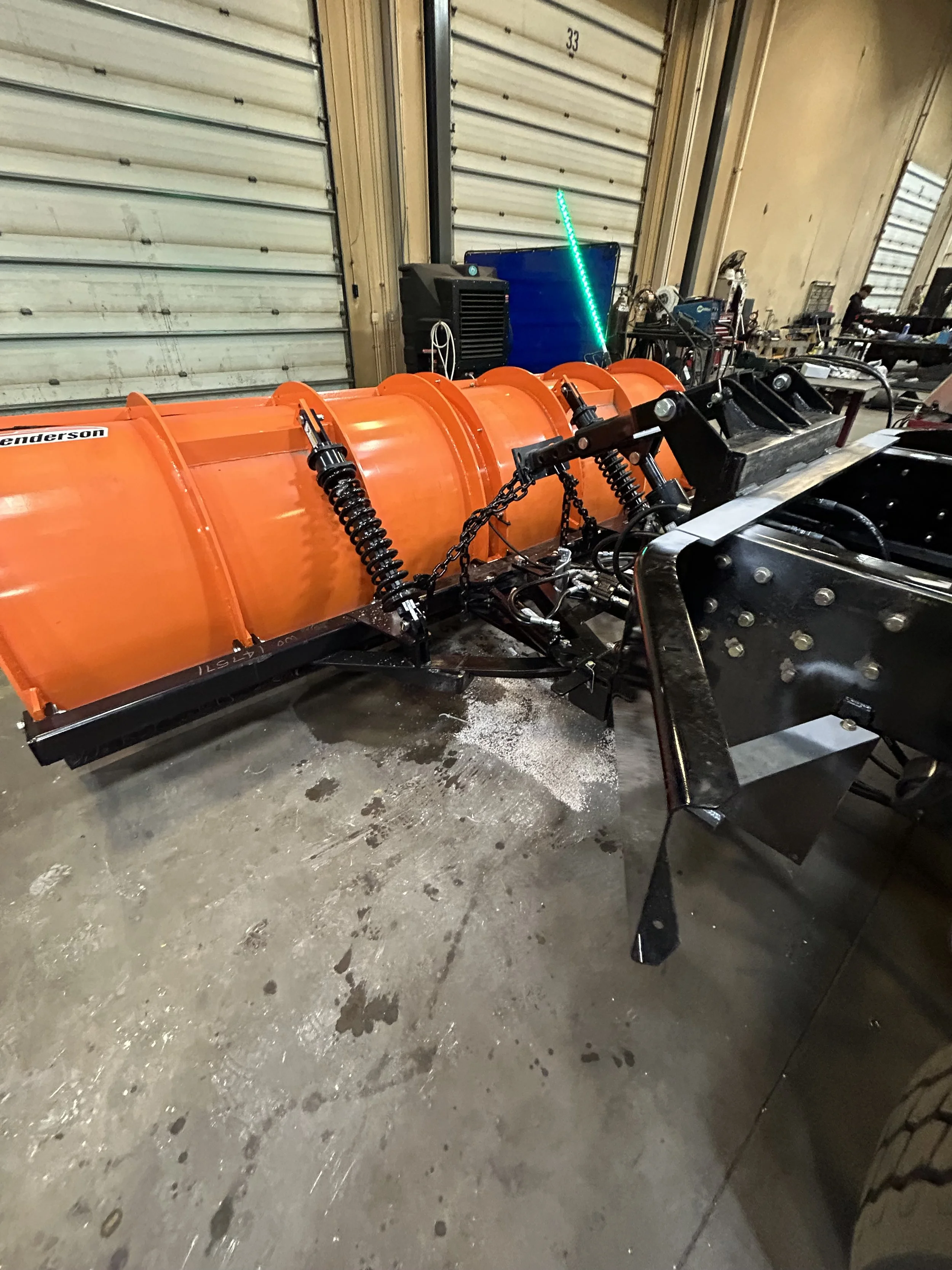 A snow plow with an orange blade, black attachment, and hydraulic shock absorbers sitting in a garage or warehouse with a concrete floor and partially open metal roll-up doors.