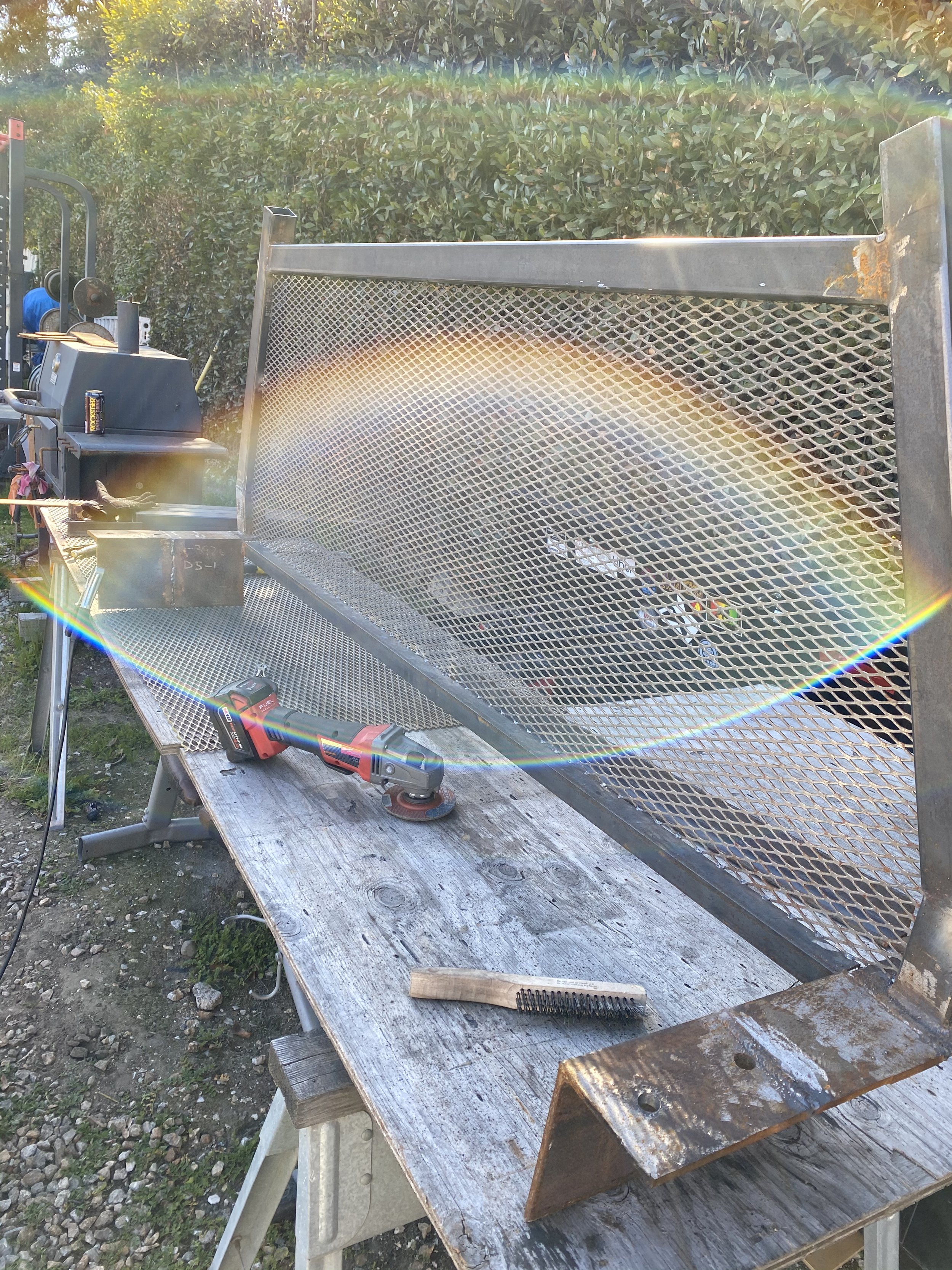 A metal workbench outdoors with a partially assembled metal frame, power tools, a pair of safety gloves, a brush, and an open bottle of water. There is a wooden table surface holding some equipment and a background of greenery and bushes.