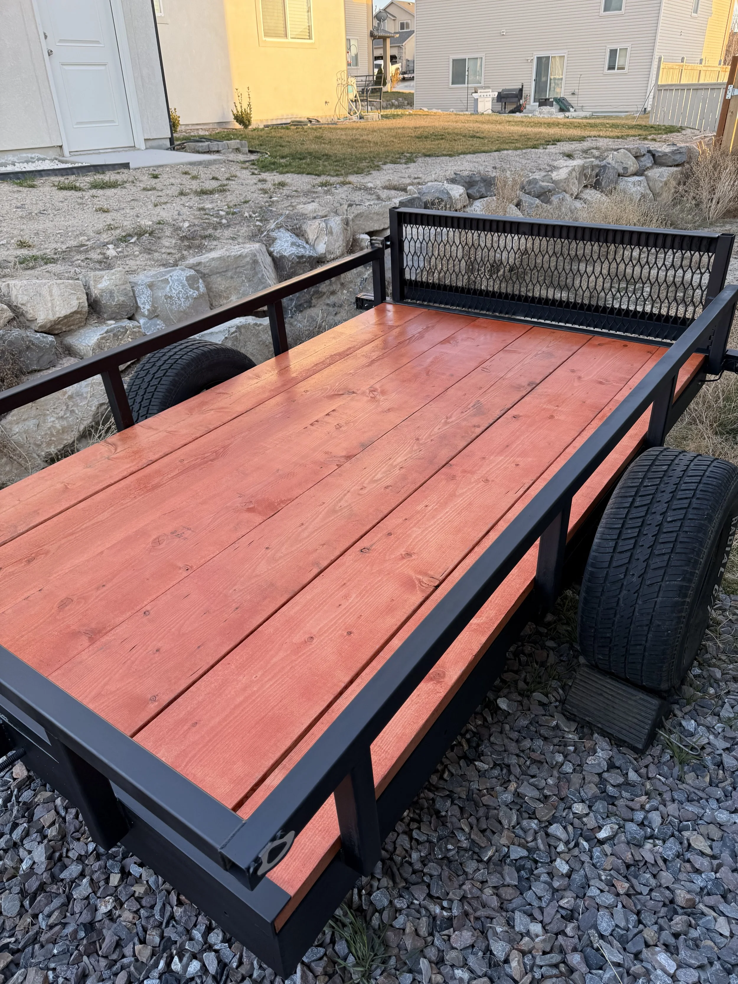 A wooden flatbed trailer with black metal frame and large tires, parked on gravel in a backyard with rocks, grass, and houses in the background.