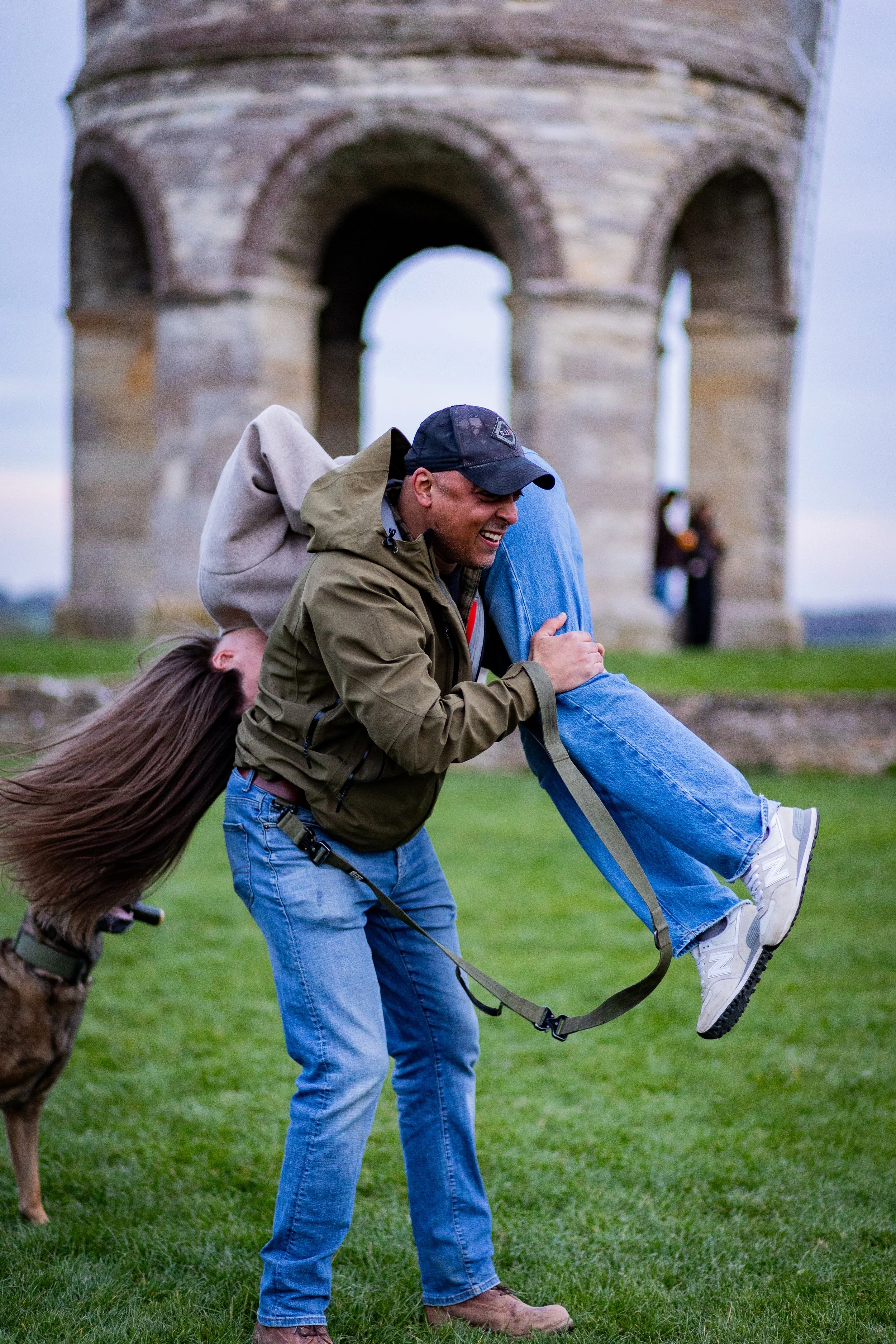 A man with a cap and sunglasses is holding another person upside down. A dog is standing nearby, and the background features an old stone arch structure on a grassy field.