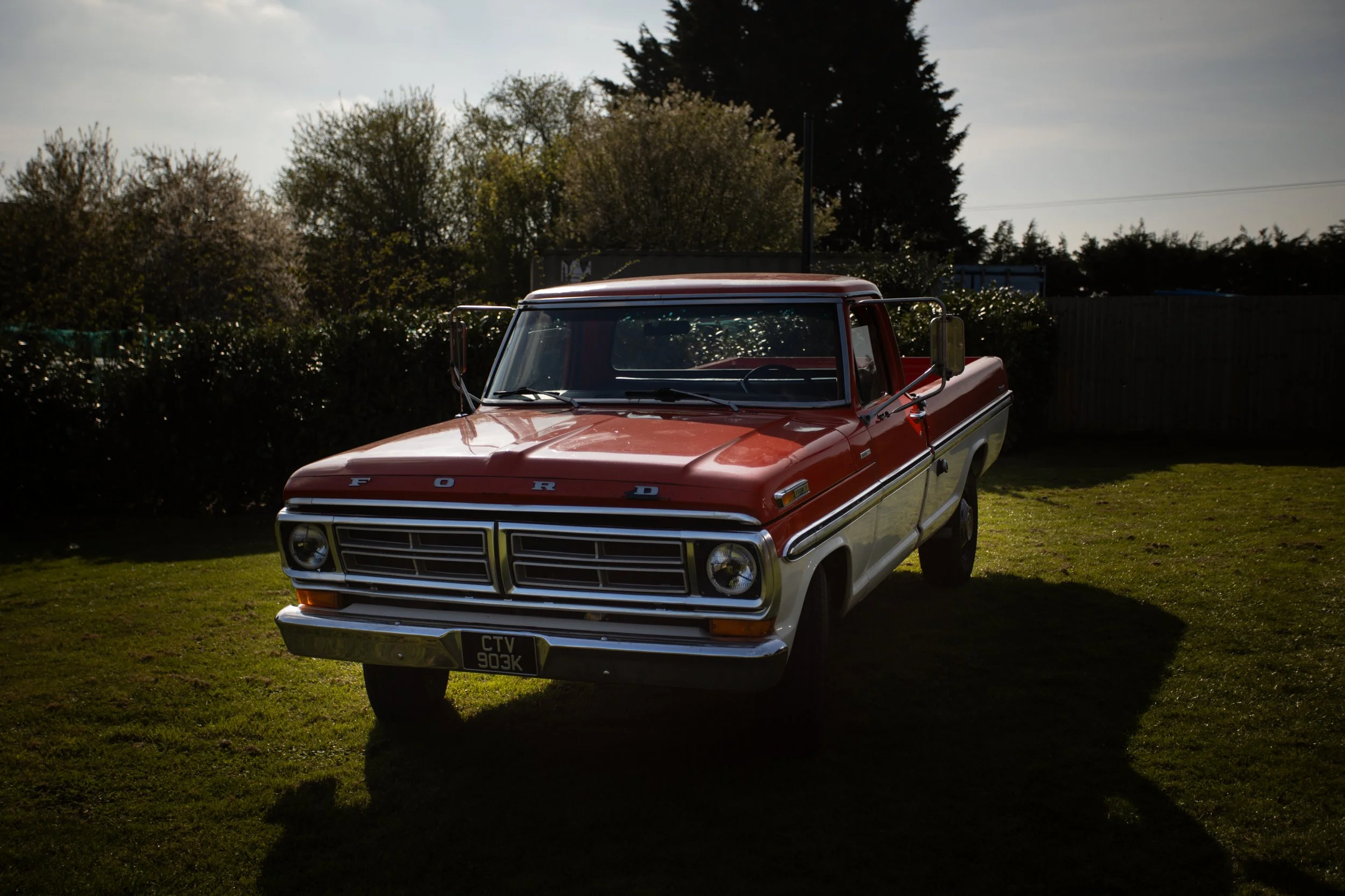 Red and white vintage Ford pickup truck parked on grass with trees and partly cloudy sky in the background.