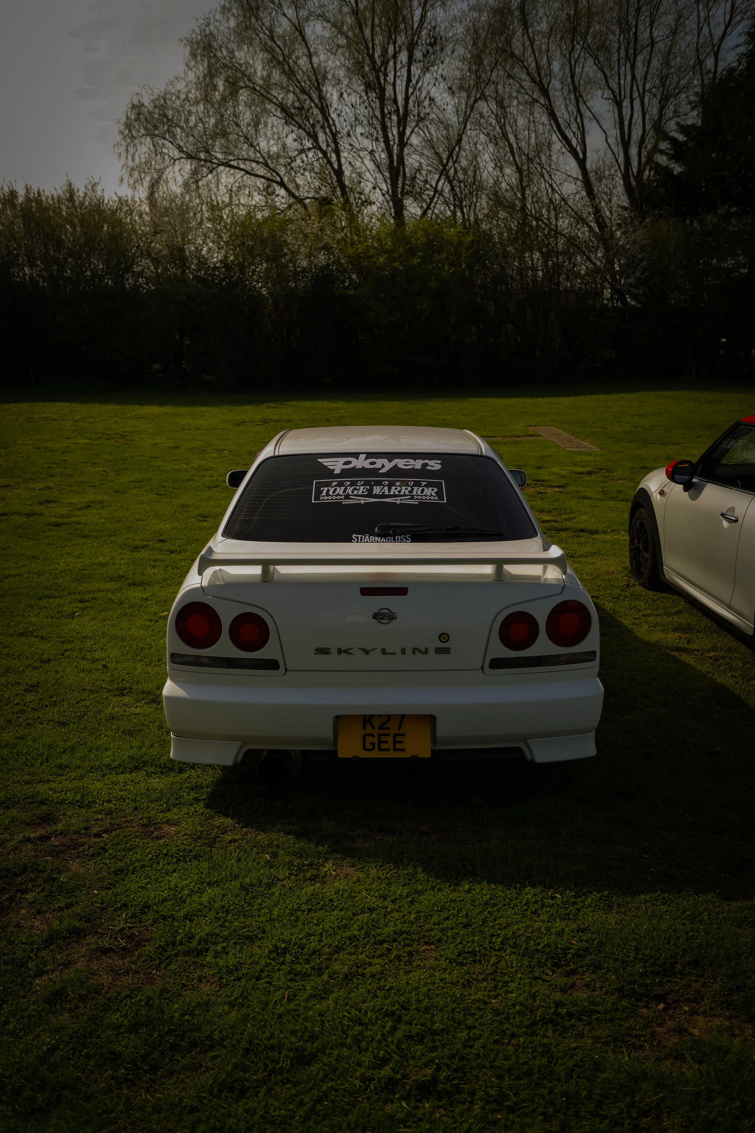 White Nissan Skyline car parked on grass, with stickers on the rear window, including 'Players,' 'TOUGE WARRIOR,' and 'STARNAGLOSS,' and a yellow license plate reading 'K27 GEE.'