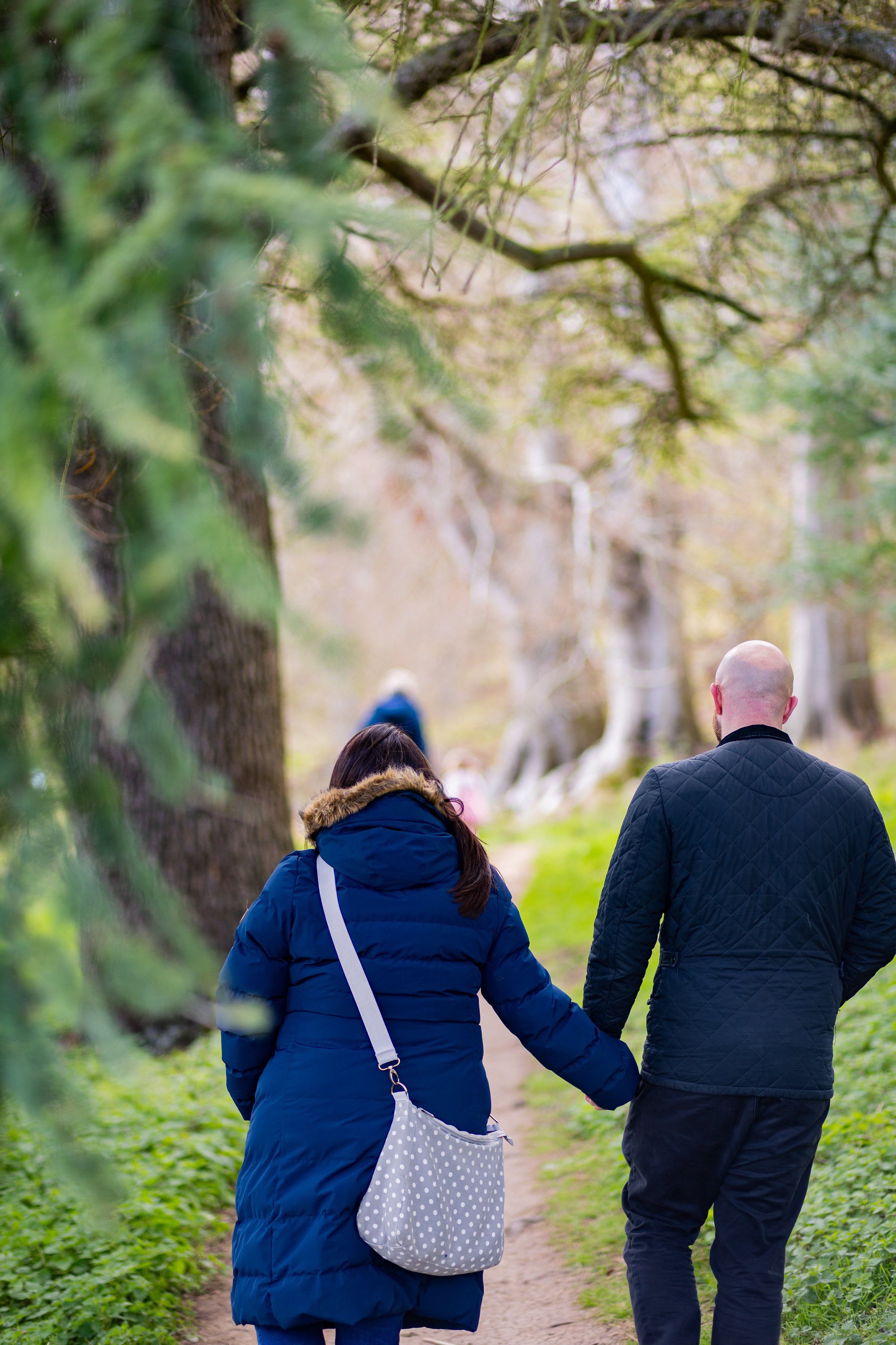 A couple holding hands walking down a wooded trail in a park with other people in the distance, surrounded by trees and greenery.
