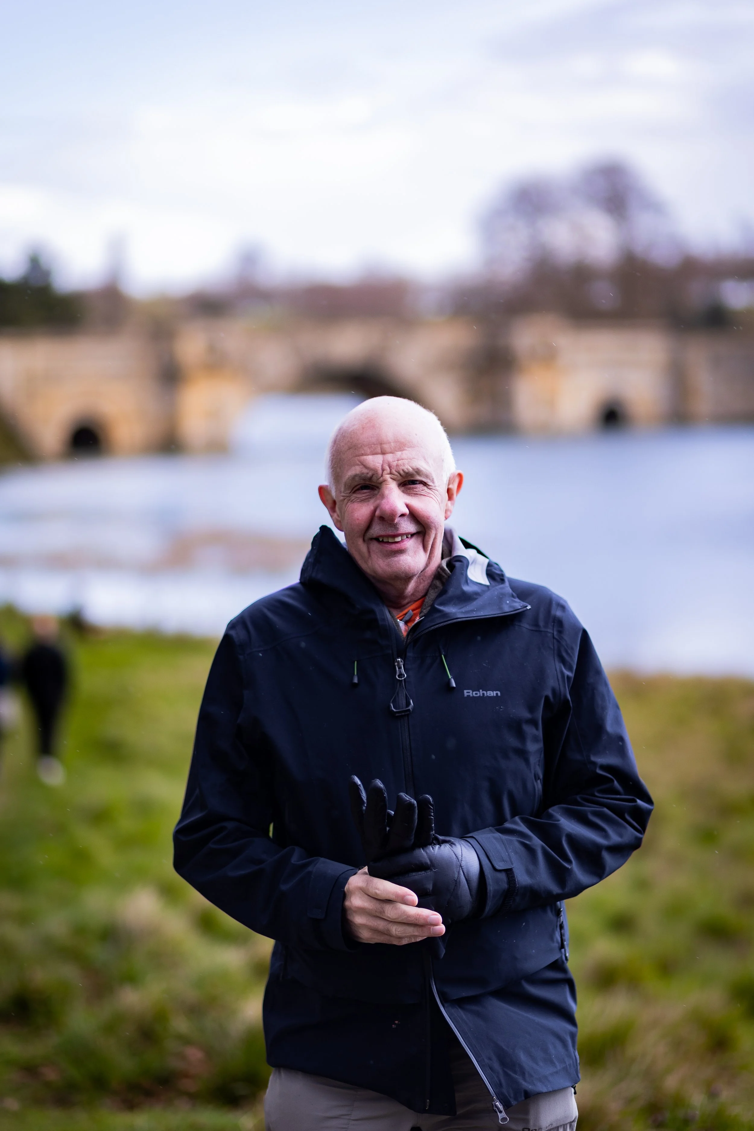 A smiling elderly man wearing a black waterproof jacket and gloves, standing outdoors near a river with a historic stone bridge in the background, on a cloudy day.