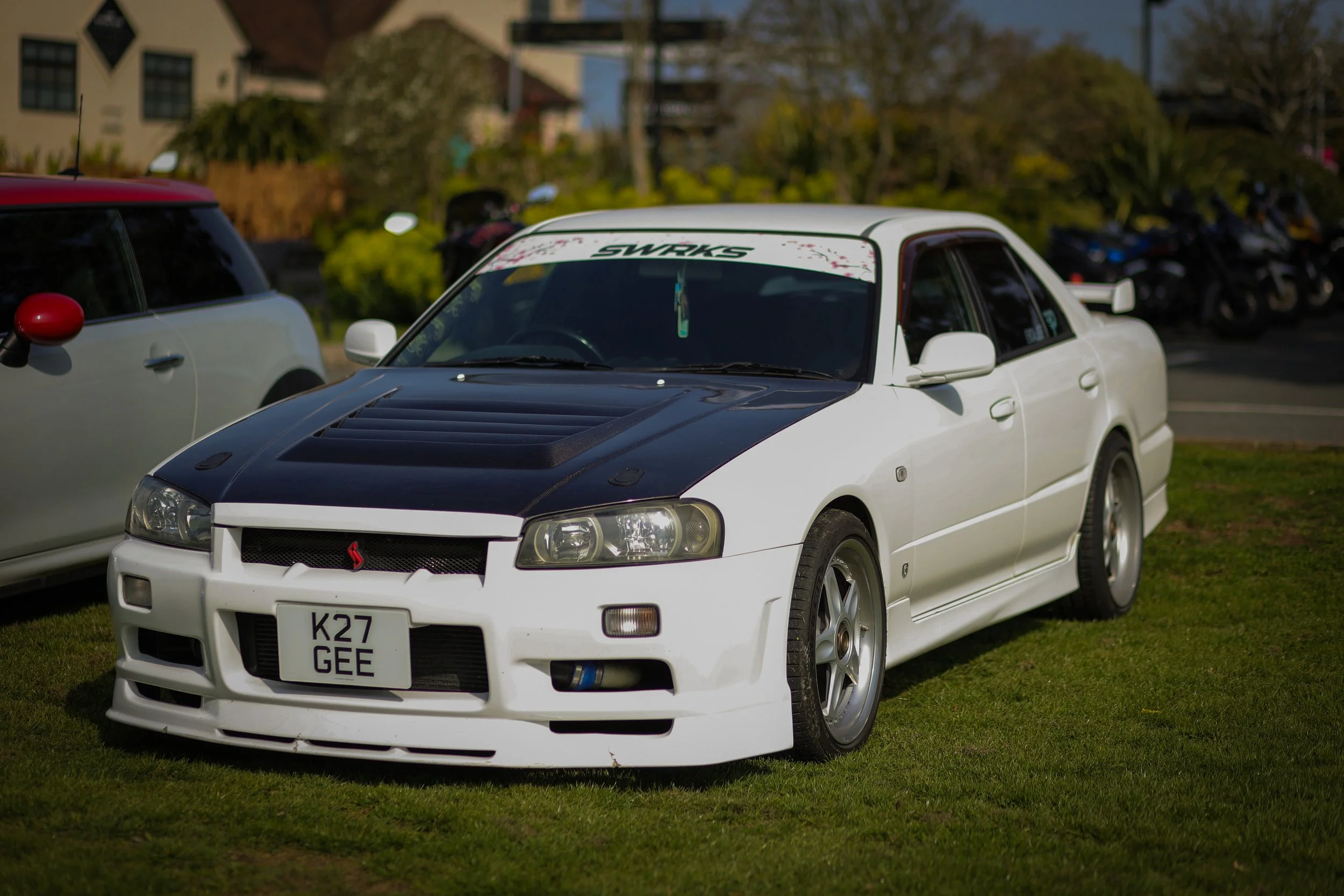 White sports sedan with black hood and custom wheels parked on grass at a car show, with other cars and trees in the background.