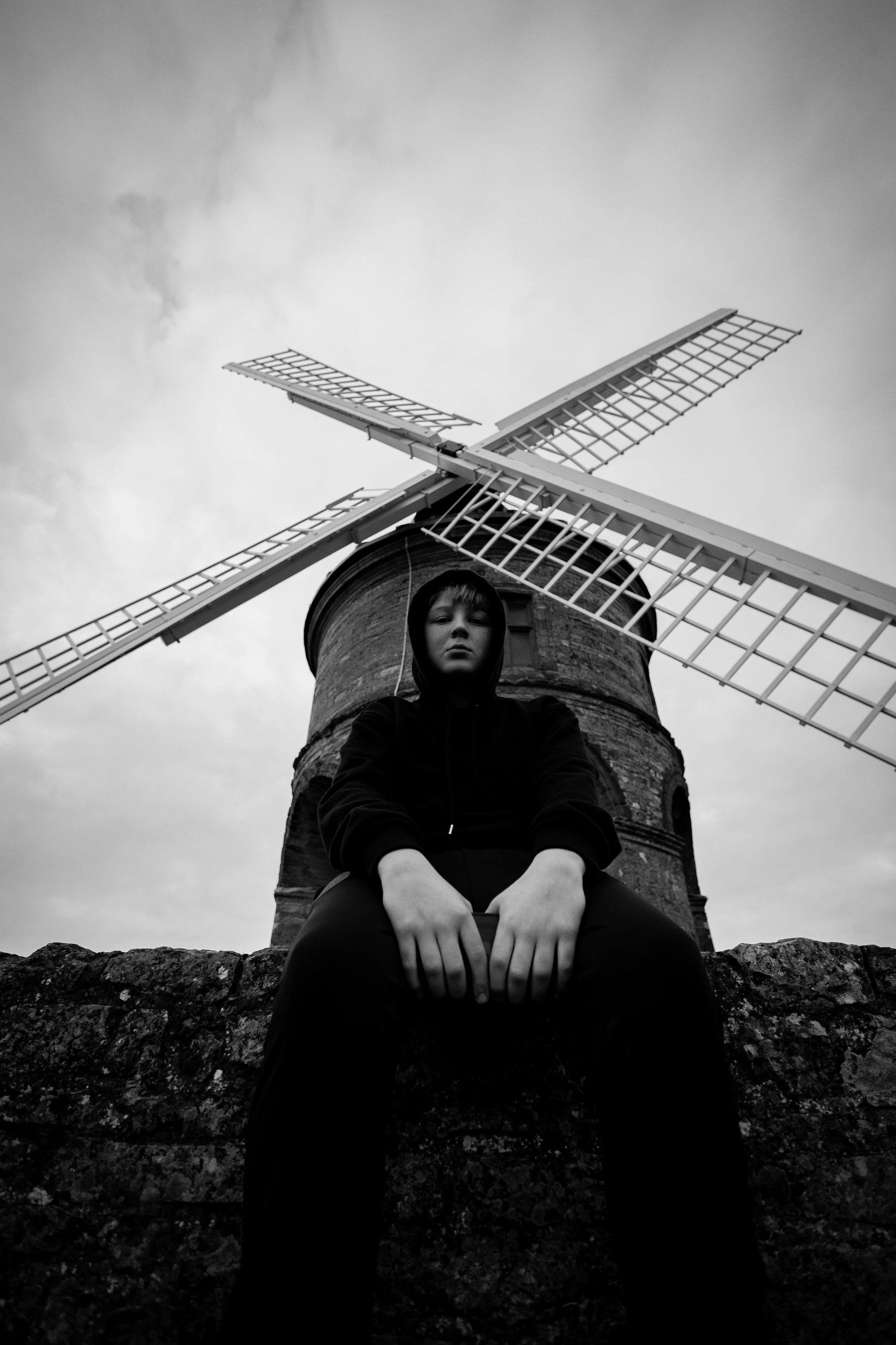 A black and white photo of a young person sitting on a stone wall in front of a windmill, with clouds in the sky above.