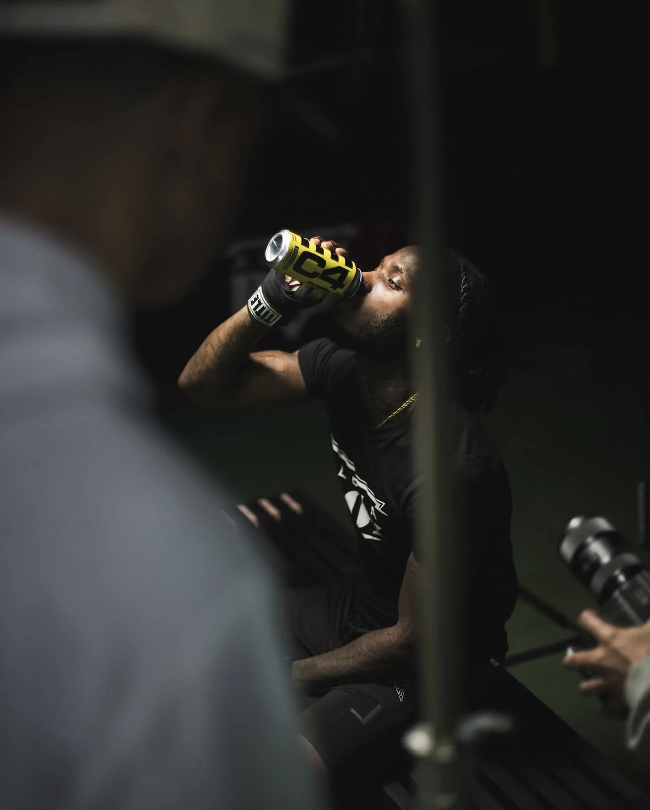 A man with long hair and a black shirt is sitting and drinking from a yellow and black can, surrounded by gym equipment in a dimly lit gym.