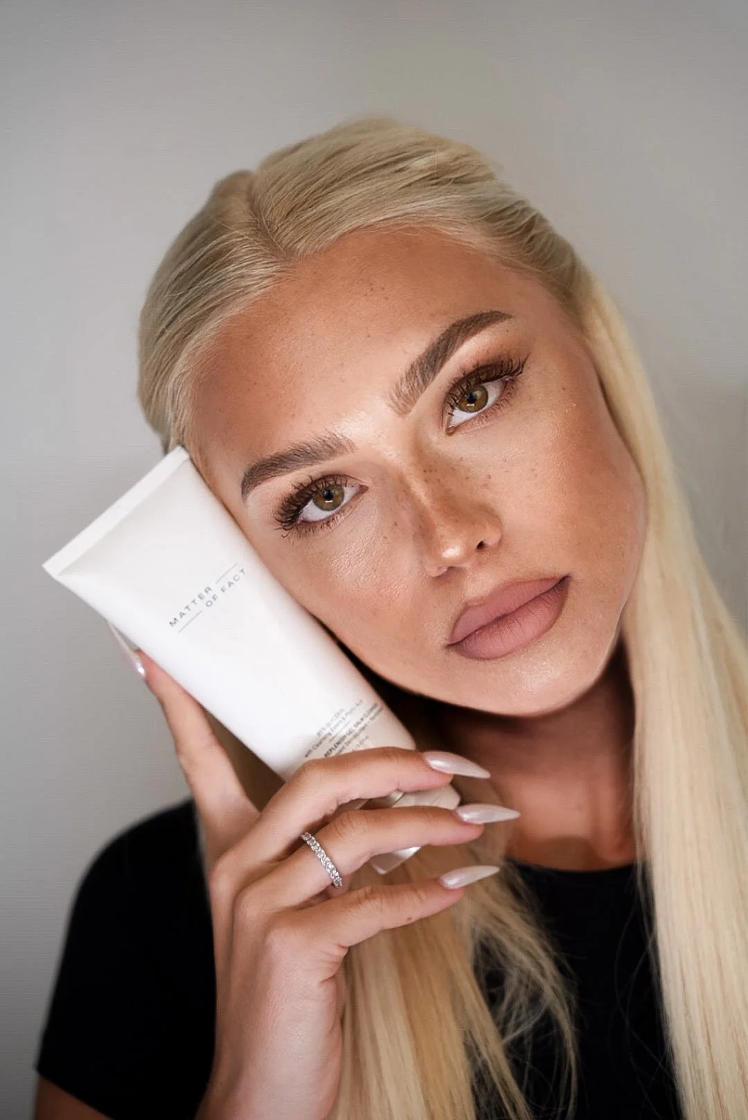 A woman with blonde hair and makeup holding a white skincare tube against her face.