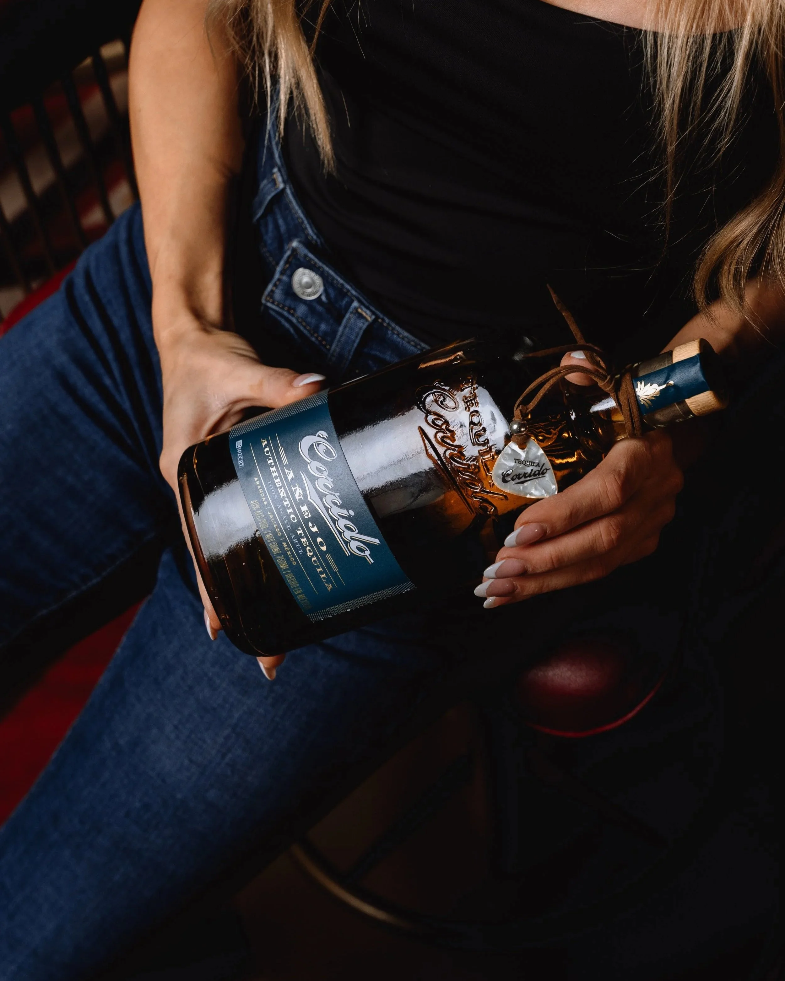 Person holding a bottle of tequila, sitting with legs crossed, wearing a black top and blue jeans, with a red and black background.