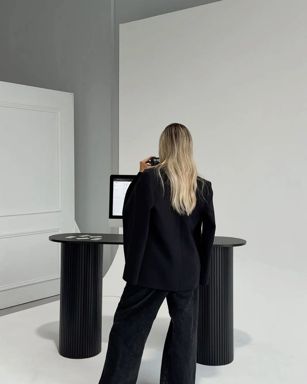 A woman with long blond hair, dressed in a black blazer and pants, stands at a display table looking at a monitor and holding a phone in a minimalistic setting.