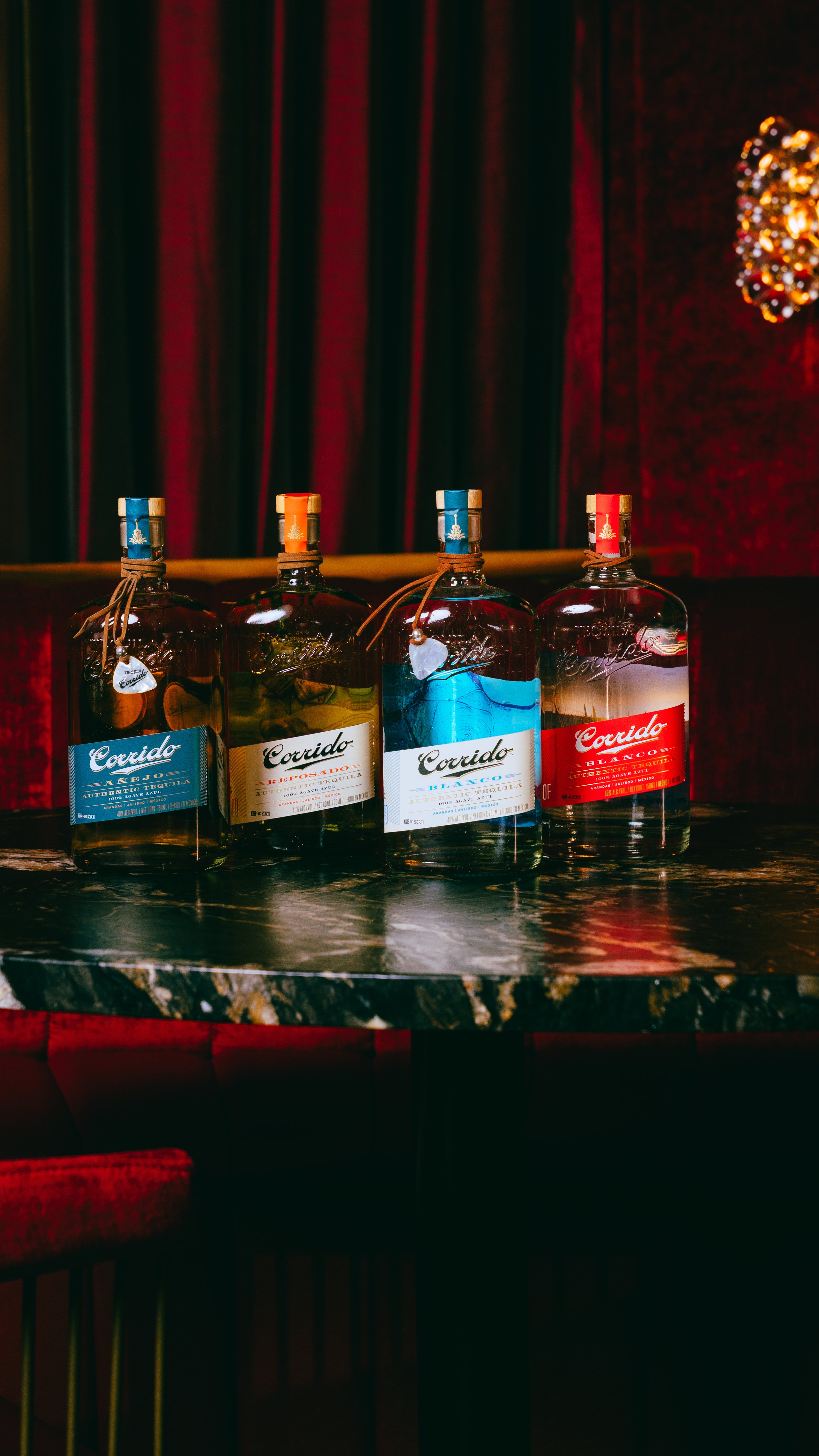 Four bottles of tequila on a dark marble table in front of a red velvet curtain, with a Holiday season decoration hanging from the ceiling.