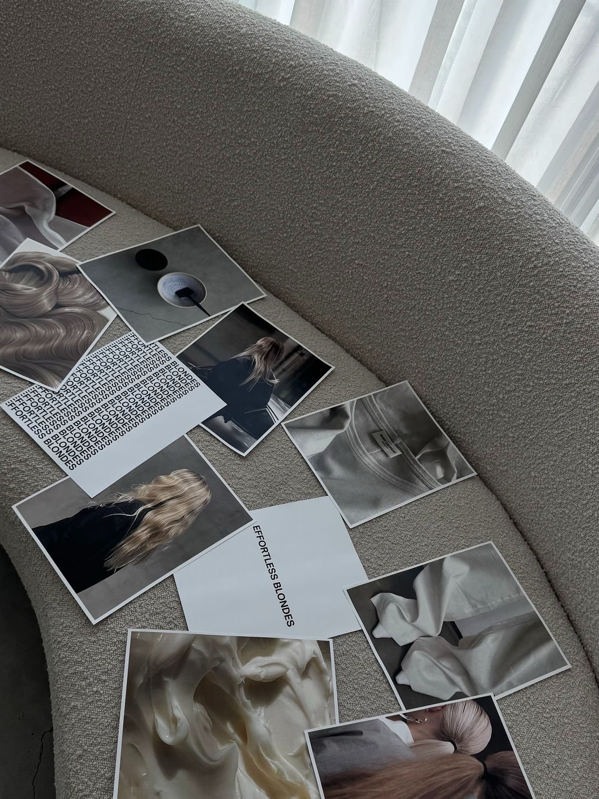 Multiple printed photographs and pieces of paper featuring images of blonde hair, a woman sitting on a bench, fabric textures, and monochrome patterns spread out on a beige fabric surface near a window with sheer curtains.