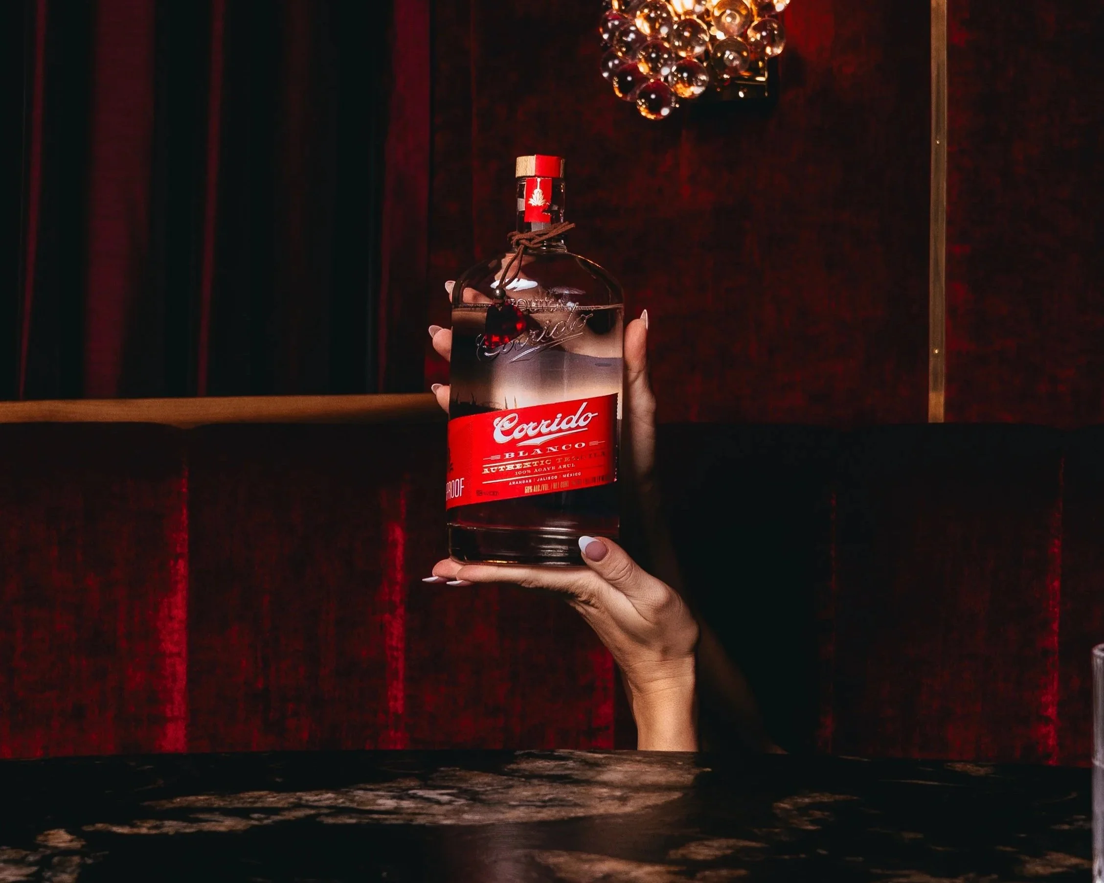 A hand with light-colored nail polish holding a bottle of Coyido Blanco tequila against a dark, red-wood textured background with a modern chandelier overhead.
