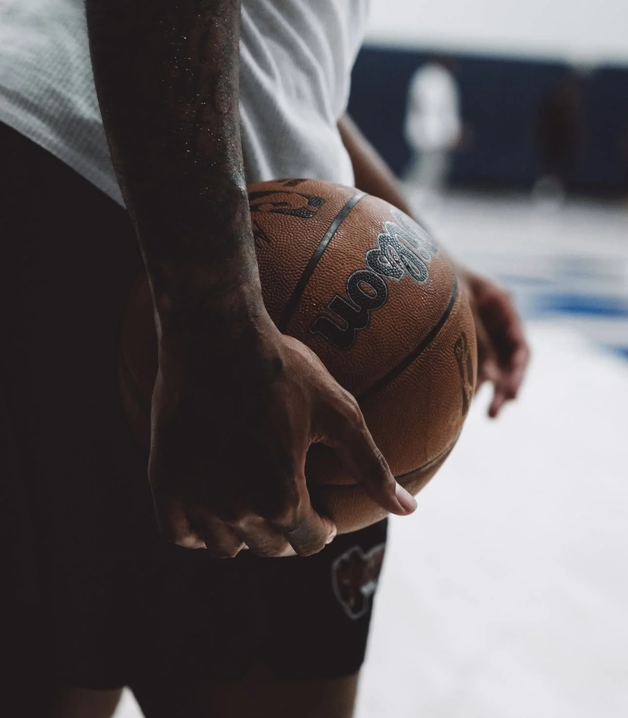 A person holding a basketball with their right hand, gripping it from the side, wearing a white shirt and black shorts with a logo, in an indoor setting.