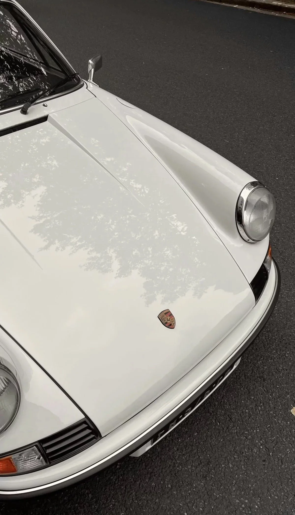 Close-up photo of a white vintage Porsche sports car, showing the front left side with a Porsche emblem on the hood and a round headlight.