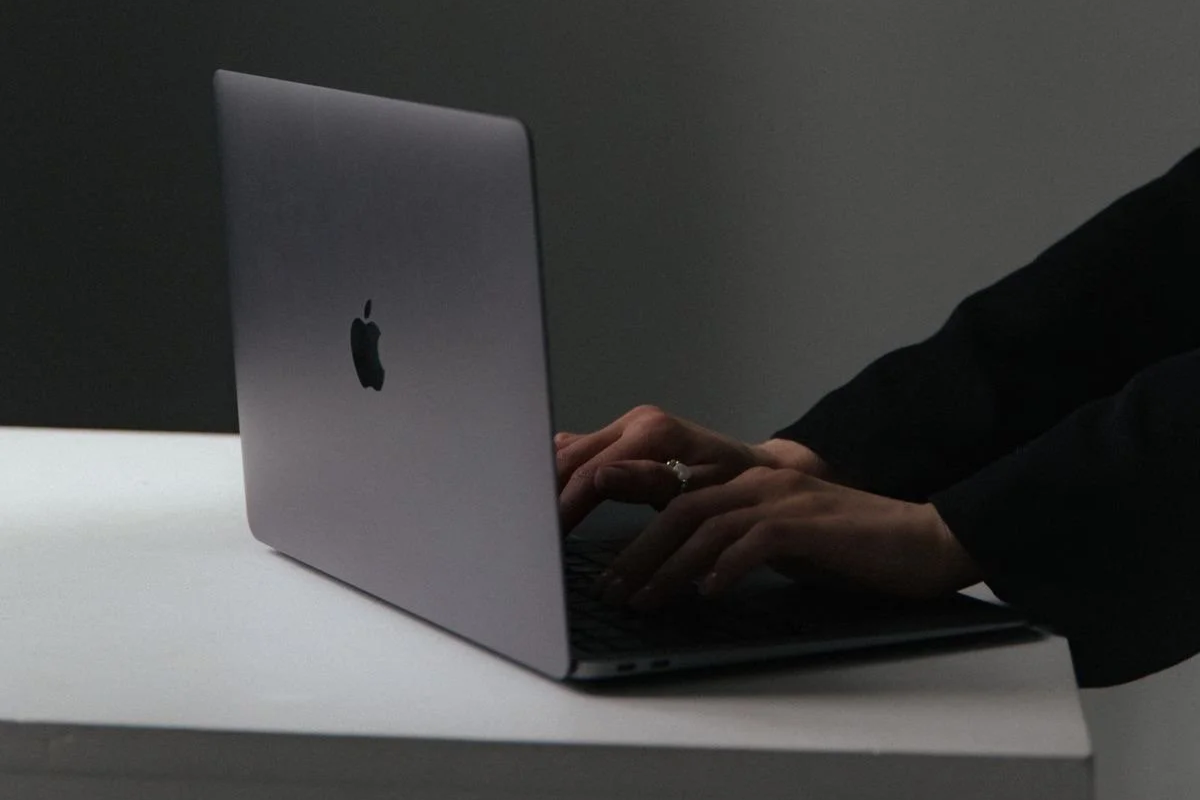 Person wearing a dark blazer typing on a silver MacBook laptop on a white table.