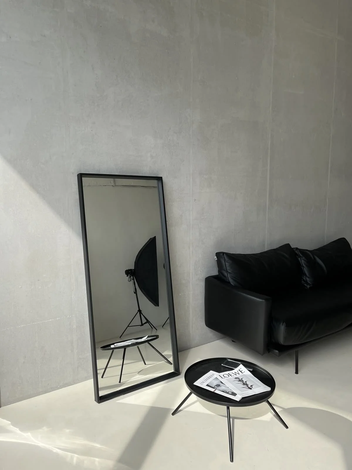 Interior of a modern room with a full-length mirror, black sofa, round black coffee table with magazines, and a studio light reflected in the mirror. The walls are gray, and the floor is white.