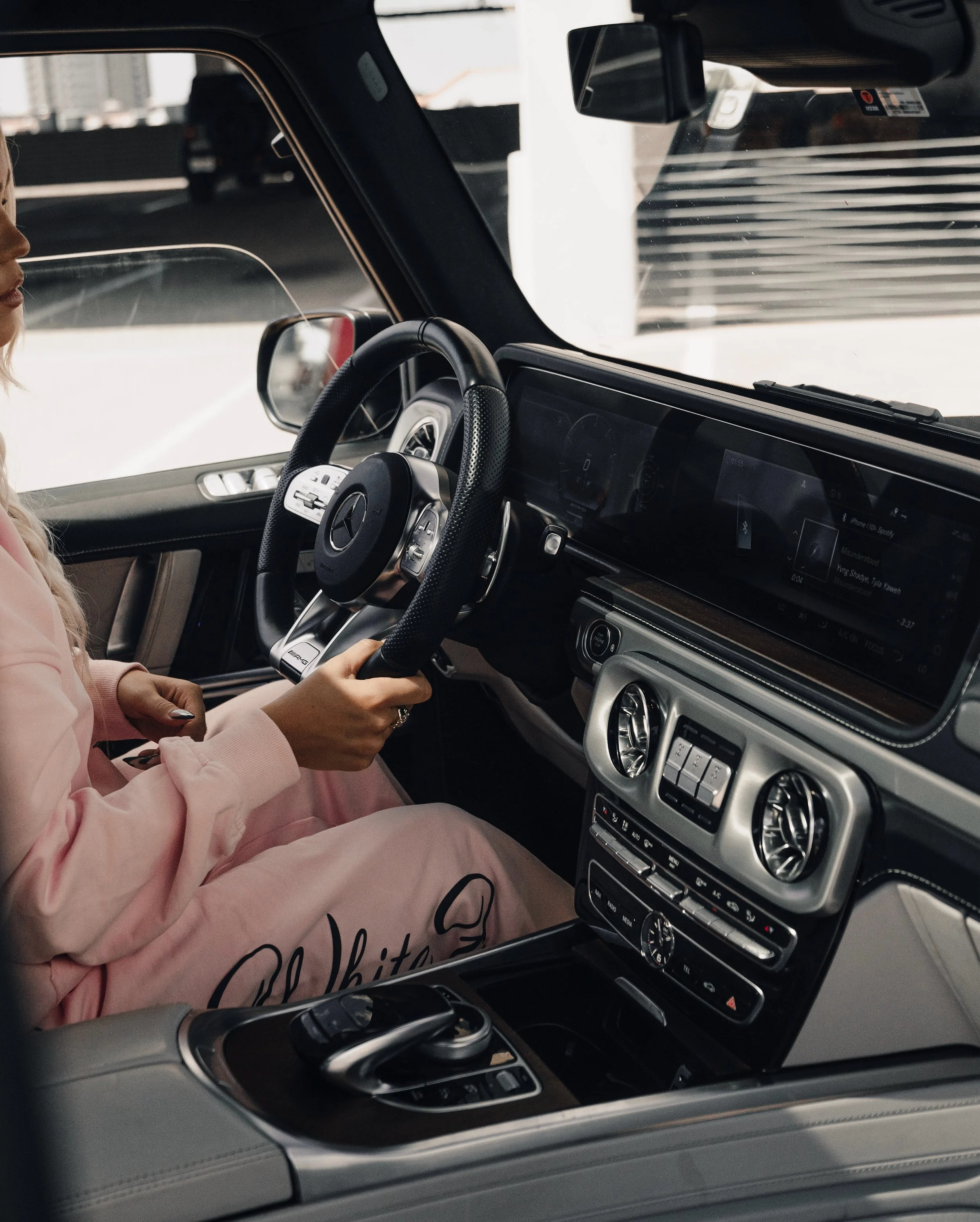 Inside of a luxury car with a woman sitting in the driver's seat wearing a pink hoodie, holding the steering wheel. The dashboard and center console are visible, featuring digital displays and control buttons. The view outside shows a city street and bridges.