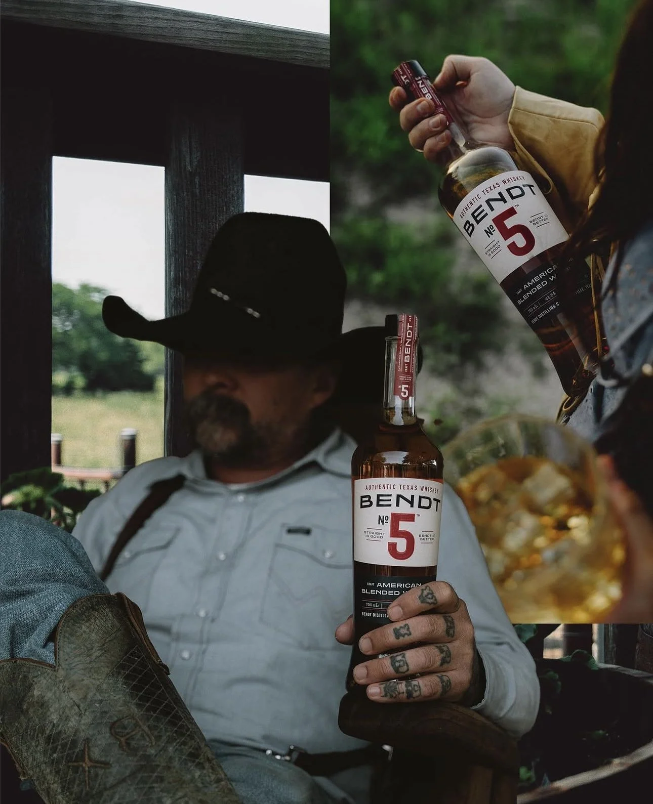 A man wearing a cowboy hat and a white shirt with suspenders sitting outdoors, holding a bottle of Bendt No. 5 whiskey, with another person holding a similar bottle partially visible. There are glasses with drinks and some food in the foreground, and greenery in the background.