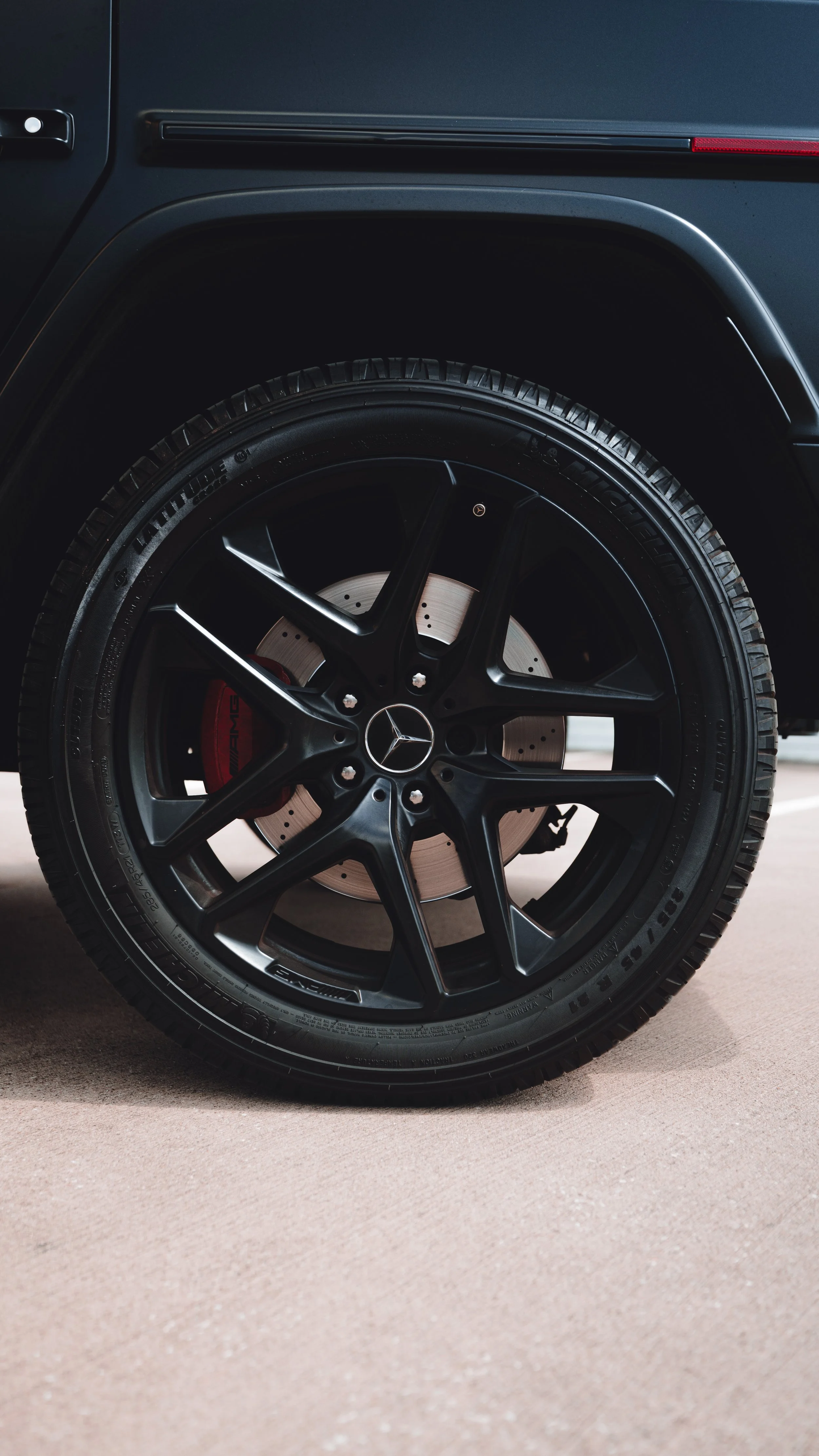Close-up of a black Mercedes-Benz car wheel with AMG brake caliper, tire, and hubcap, parked on a concrete surface.