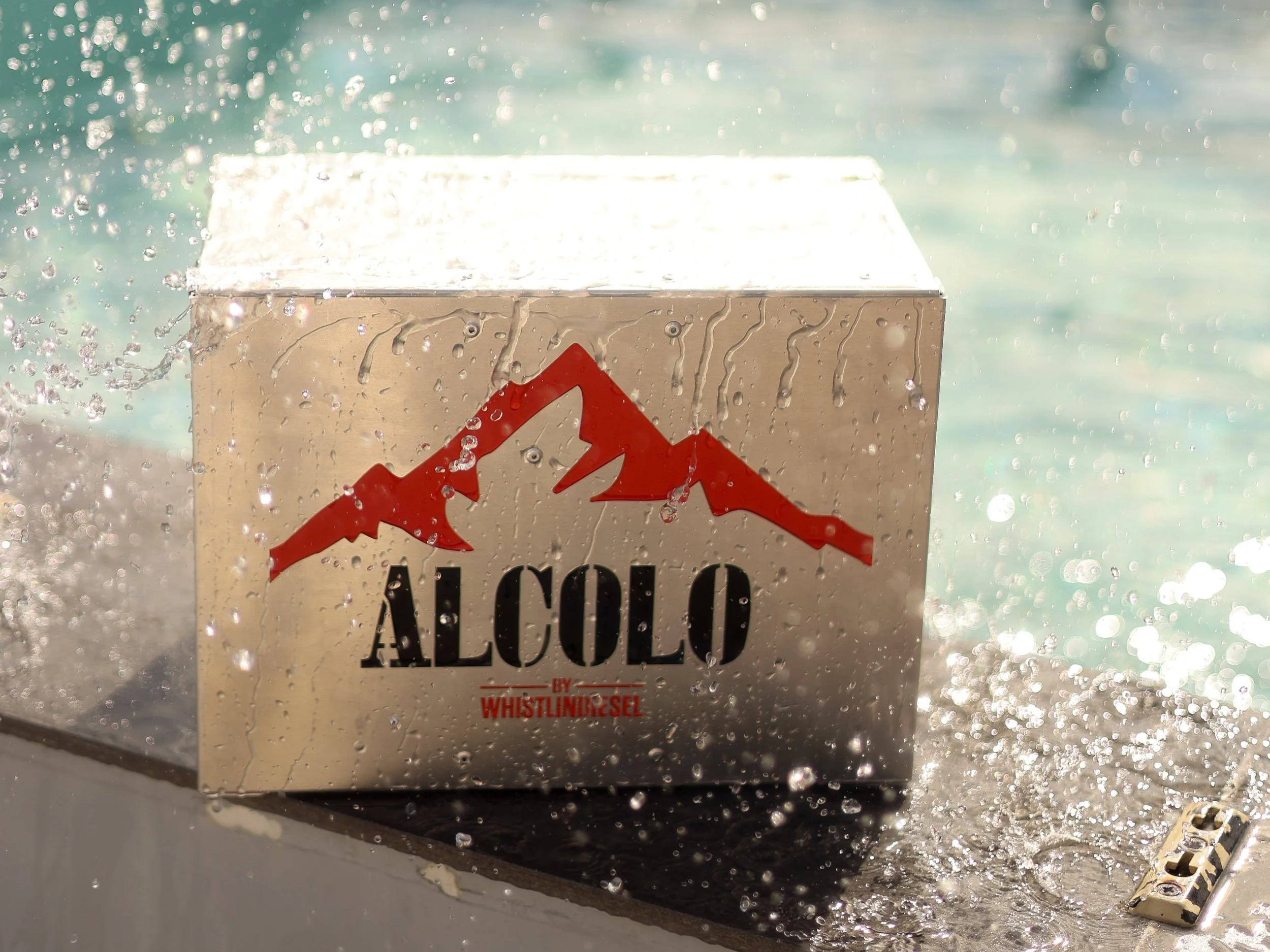 A close-up photograph of a warning sign on a metal object, with water droplets on the surface, indicating splashing or moisture. The sign features a red mountain symbol and the words 'ALCOHOL by WHISTLINBIRSEEL' beneath it.