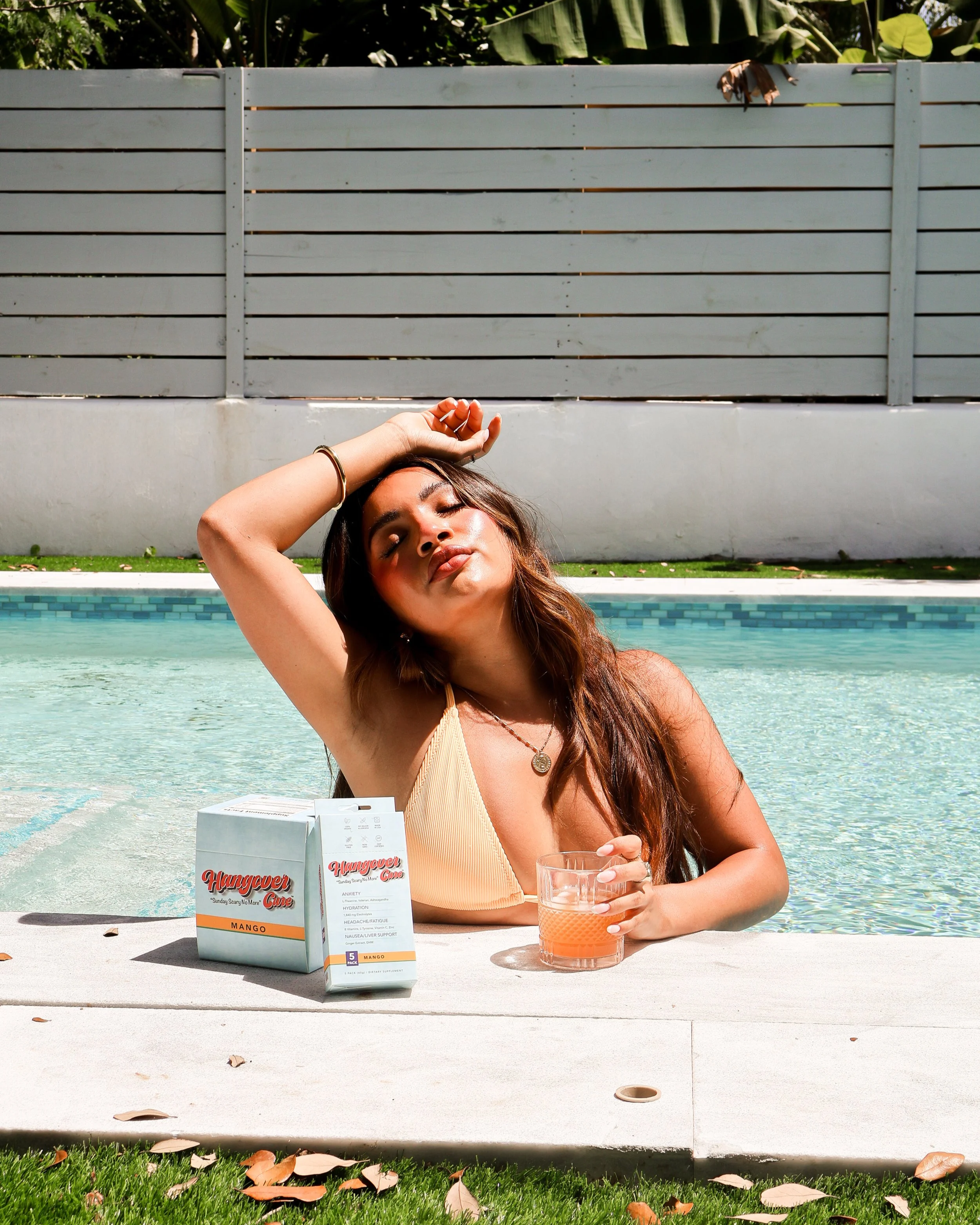 A woman with long brown hair and a beige bikini top sitting by a pool during daytime, with her eyes closed and one hand resting on her head. In front of her on the table are a box of Hangover Cure supplements labeled Mango, a glass of orange-colored beverage, and some leaves scattered on the ground.