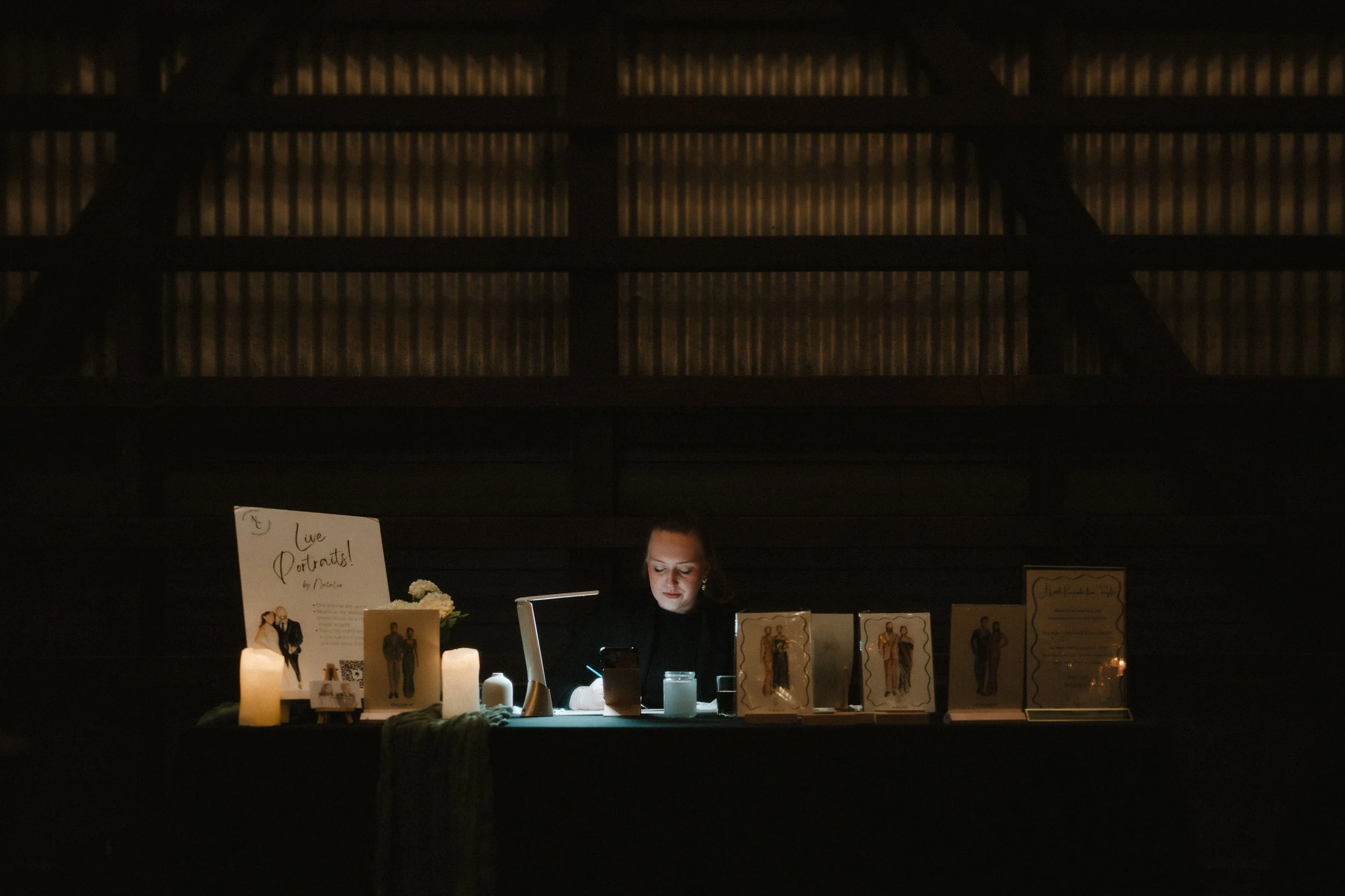 A woman sitting at a table with framed portraits, candles, and a sign that reads 'Live Portraits!' in a dimly lit room.