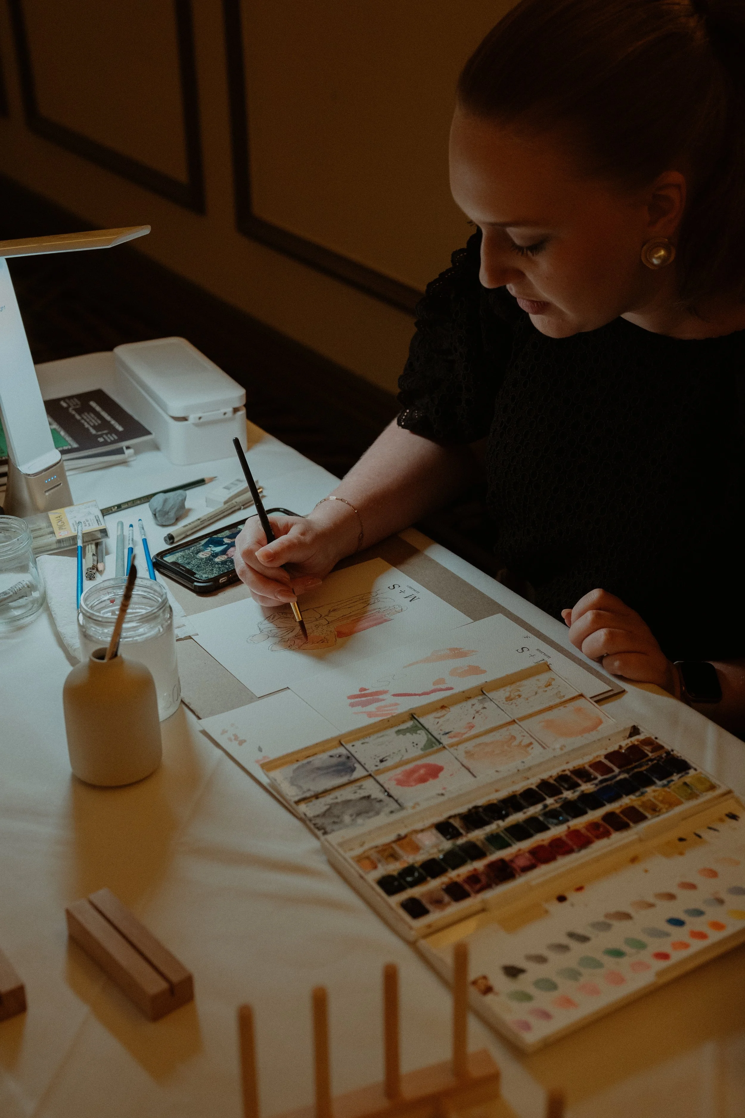 A woman sitting at a table with watercolor paints, brushes, and a palette, creating a painting or illustration.