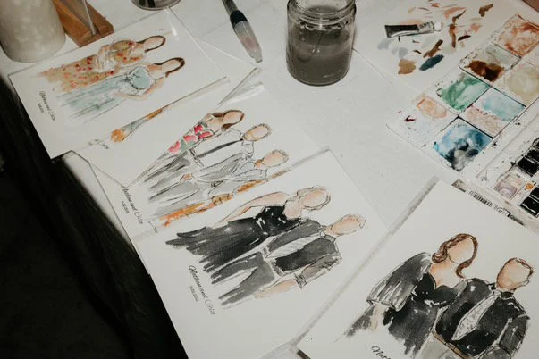 Watercolor fashion illustrations of women and men on paper, surrounded by watercolor paints, brushes, and a jar of water on a table.
