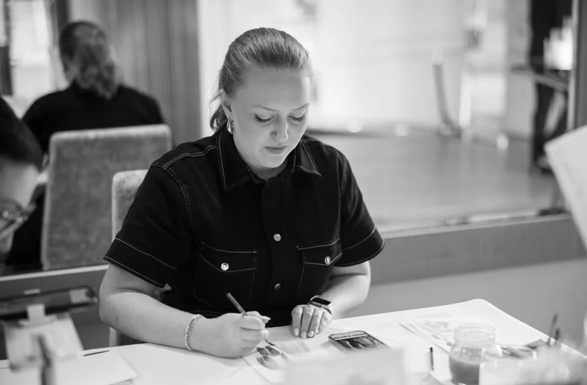 A woman sitting at a table, working with photographs and a pen, in a room with other people in the background.
