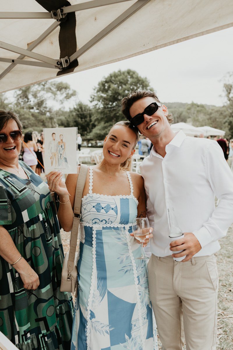 Three people smiling at an outdoor gathering, with the woman in the middle holding a painting and a glass of rosé, and the man on the right holding a bottle.