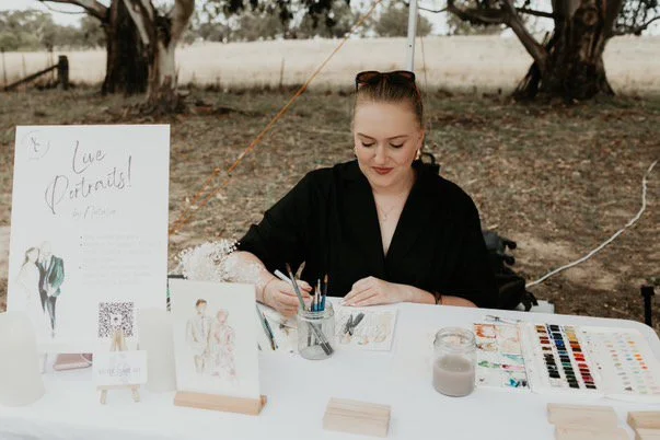 A woman sitting at an outdoor art booth, painting or drawing portraits, with art supplies on the table and a sign that reads 'Live Portraits!'
