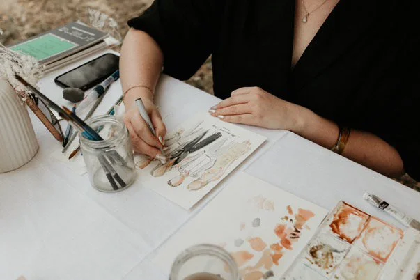 A person sits at a table with art supplies, sketching or painting in a sketchbook, surrounded by paint palettes, brushes, and water jars.