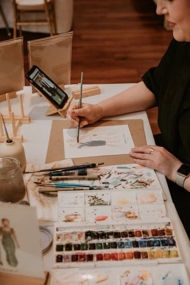 A woman sitting at a table painting a fashion illustration with watercolor paints. She is using a brush and has a smartphone propped up on a stand, displaying a reference image. The table contains various art supplies, including paint palettes, brush