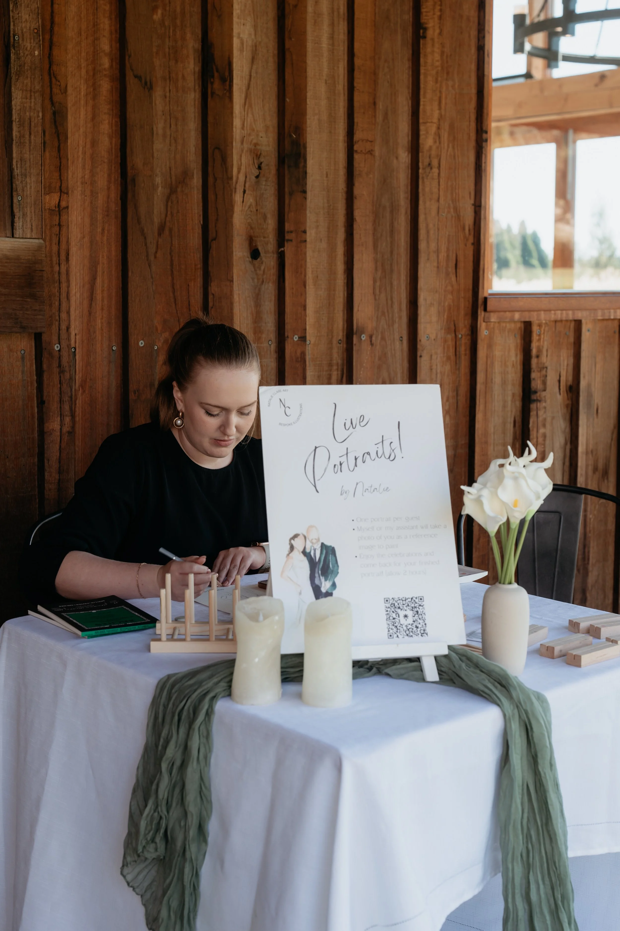 A woman sitting at a table with a sign that says "Live Portraits! by Natalie" and a sketchbook in front of her, set in a rustic wooden room with natural light, decorated with candles and flowers.