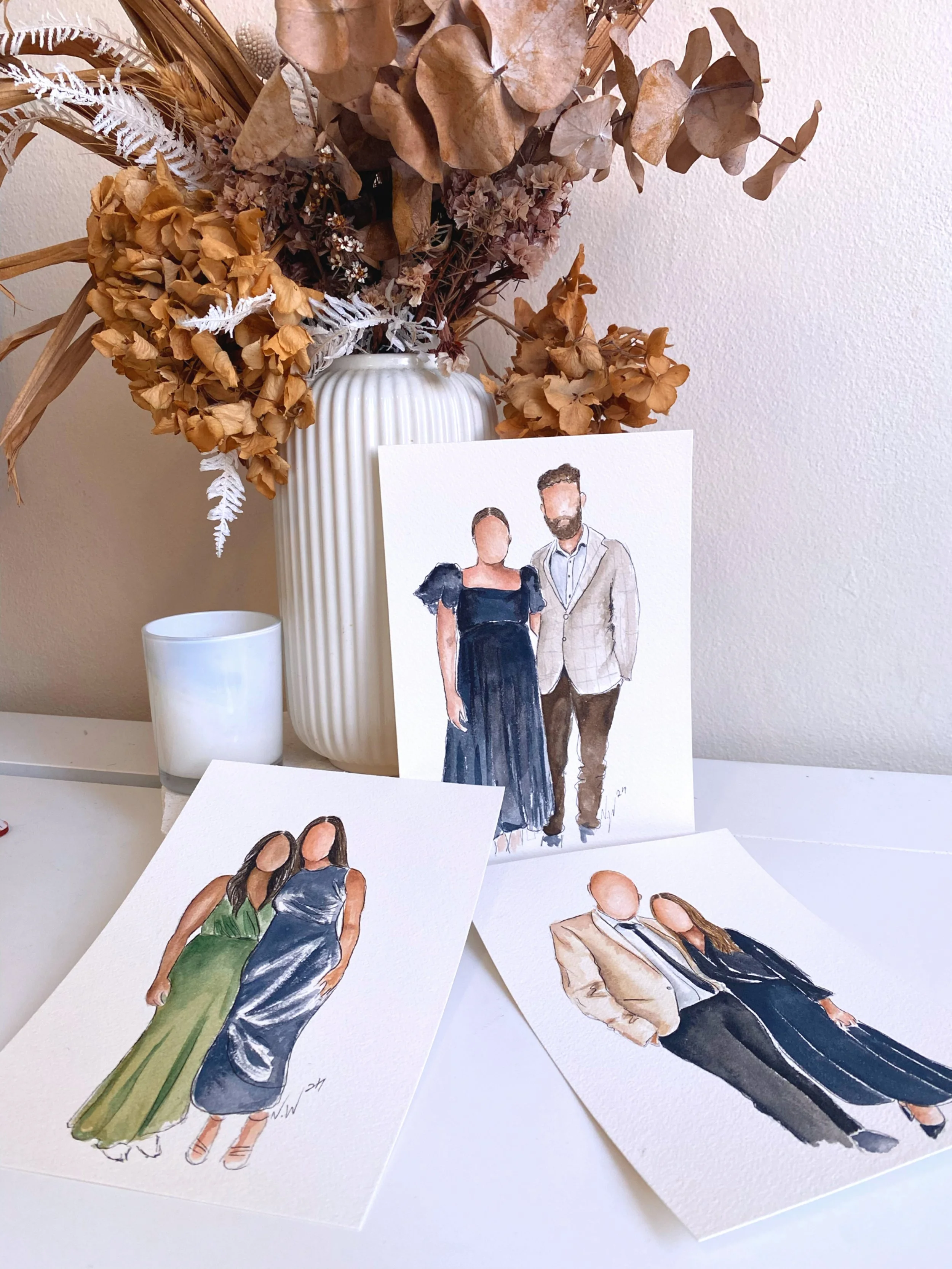 Three watercolor portraits of people in formal attire placed on a white surface, with a beige vase holding dried flowers in the background.