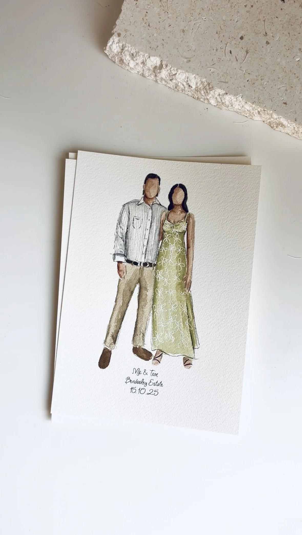 A wedding invitation featuring a watercolor illustration of a man and woman standing together, with text below reading "Me & Tom, Bendooley Estate, 15.10.25." The illustration is on textured paper, placed on a white surface with part of a beige textu