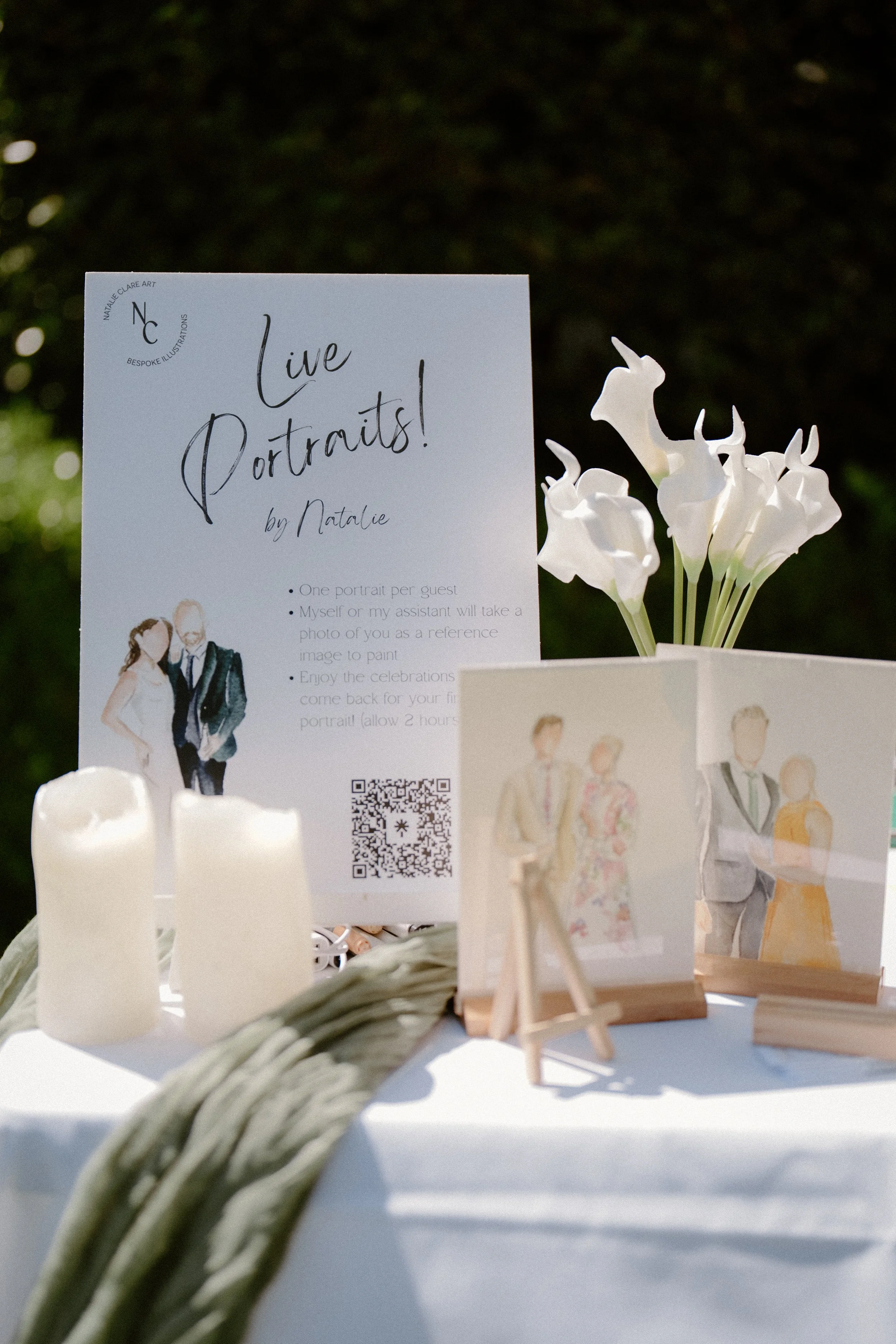 A sign advertising live portrait art at an outdoor event, accompanied by watercolor portraits, white calla lilies in a white square vase, and battery-operated candles on a tablescape.