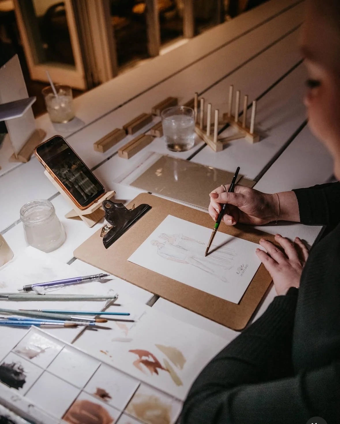A person drawing on paper with art supplies, including paintbrushes, watercolor swatches, and pencils, on a table with glasses of water and a small mirror at night.