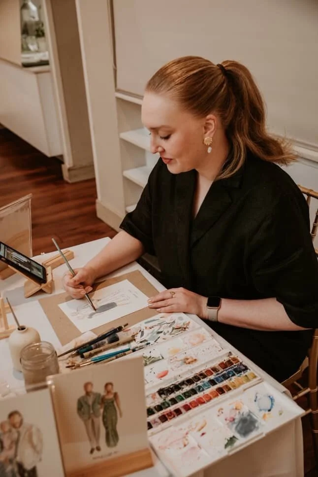 A woman with red hair tied in a ponytail, wearing a black top and earrings, is sitting at a table painting with watercolor paints. The table has art supplies, a palette, a small framed picture, and a mobile device.