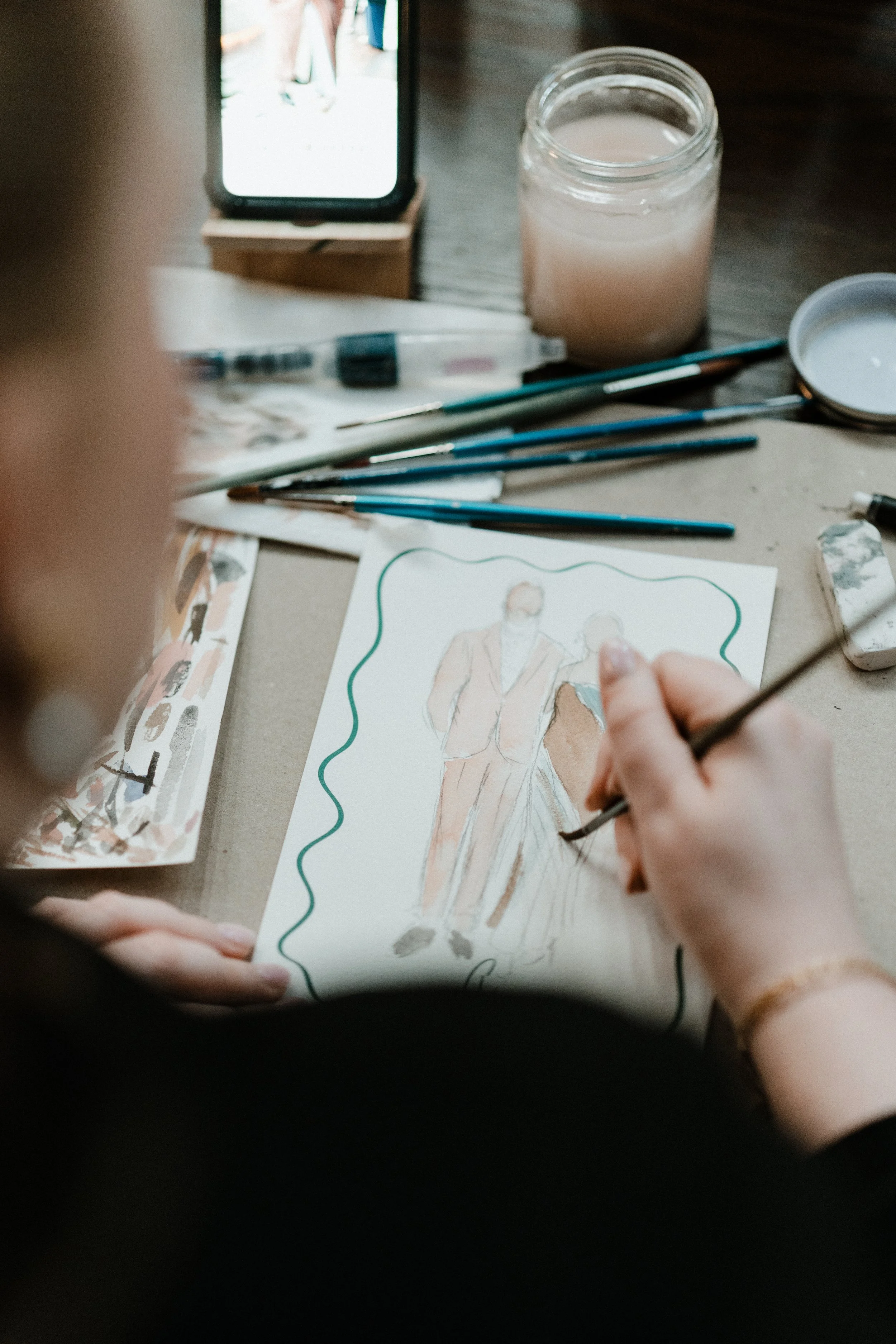 A person painting a watercolor portrait of a man and woman in formal attire, sitting at a table with various art supplies and a smartphone displaying a reference photo.