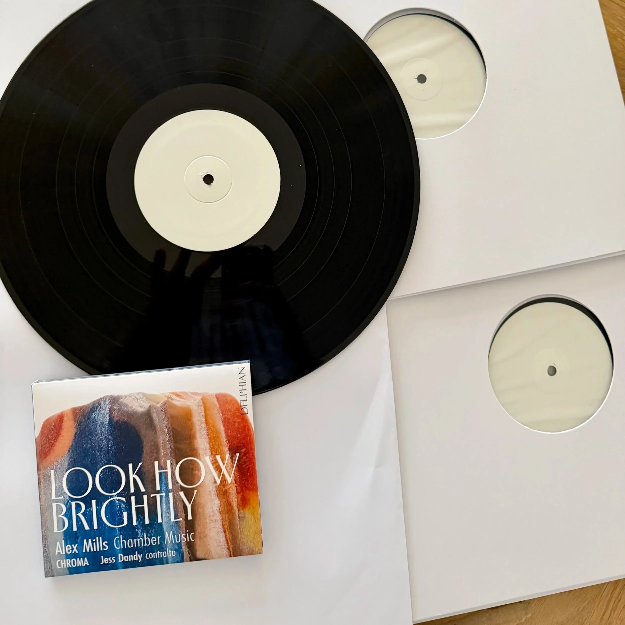 Close-up of a vinyl record with a black surface and white center label, partially visible on a white surface along with a booklet titled 'LOOK HOW BRIGHTLY' by Alex Mills with cover art by Dawn Ng