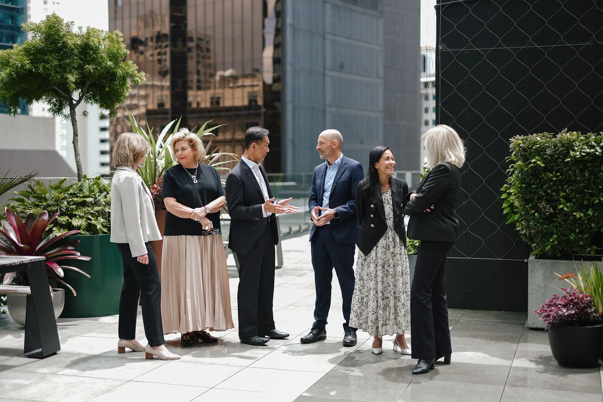 Six individuals engaged in conversation on a rooftop in an urban setting, surrounded by greenery and modern buildings.