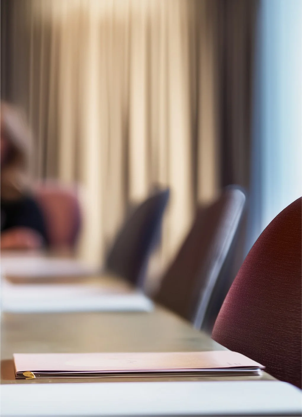 A conference room with a wooden table, pink folders, and chairs. In the background, beige curtains cover a window.